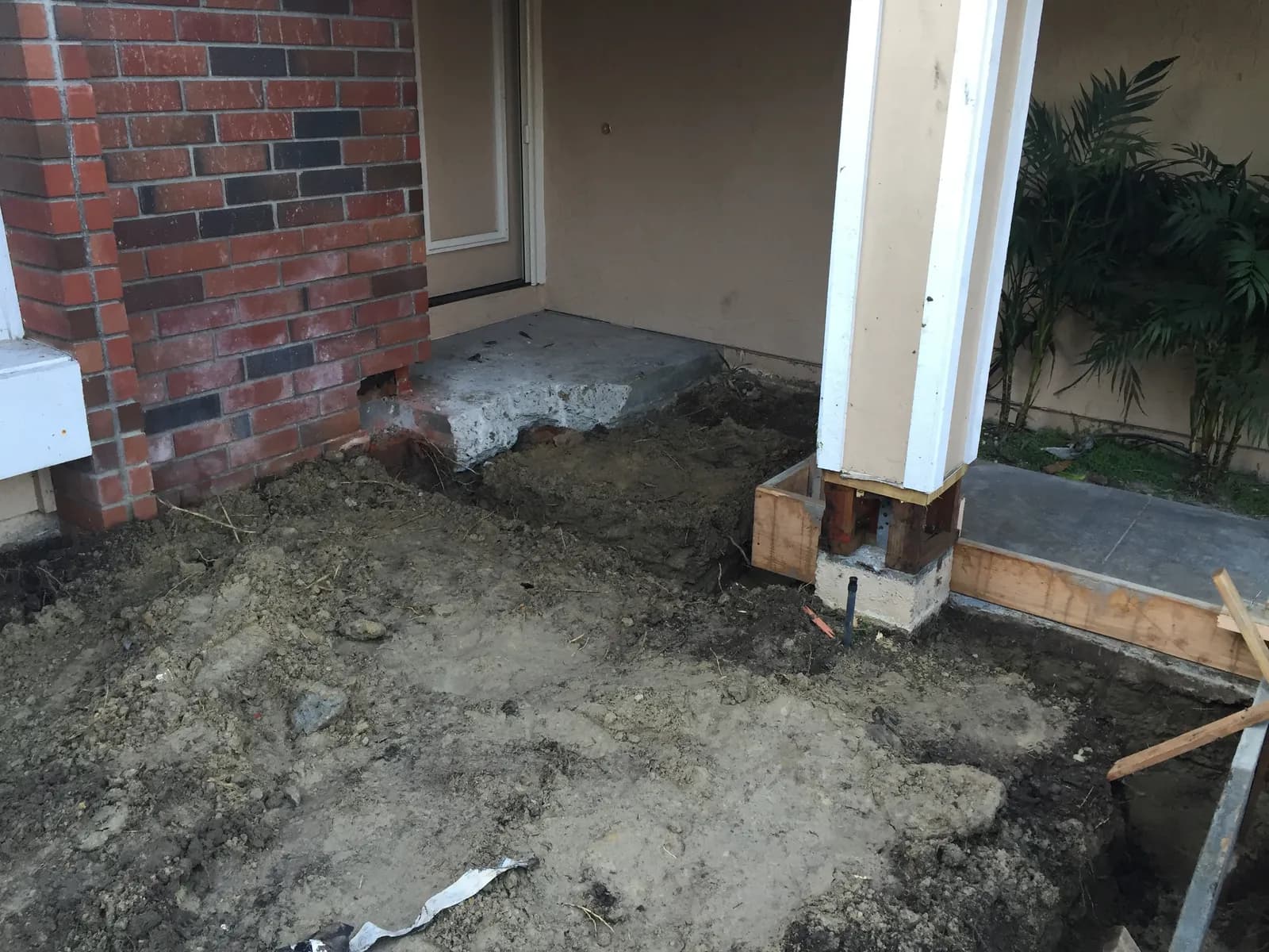 Excavated dirt and a partially demolished concrete stoop beside a brick wall and a porch post during construction.
