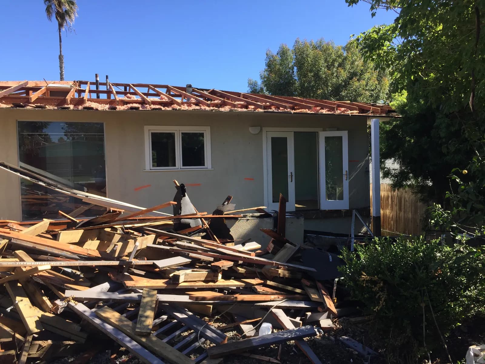 A single-story house mid-demolition with exposed roof rafters and a pile of lumber debris in the yard.