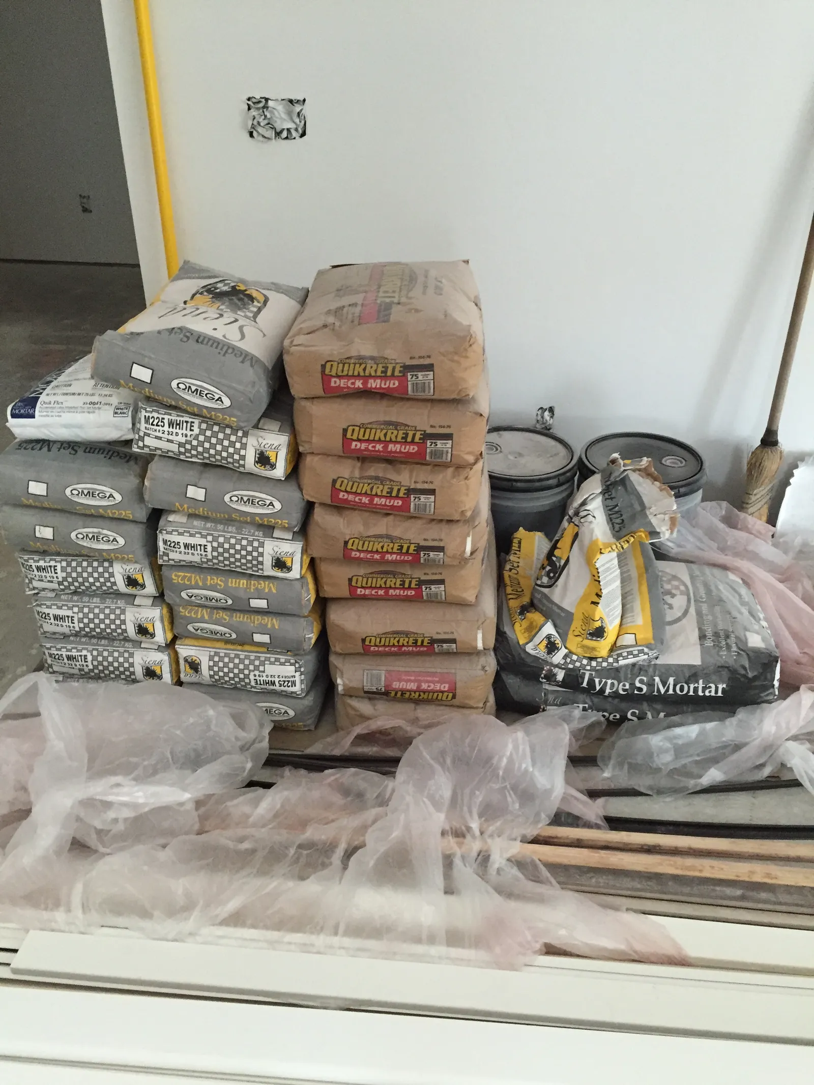Bags of mortar and deck mud stacked with buckets and tools in a room under renovation.