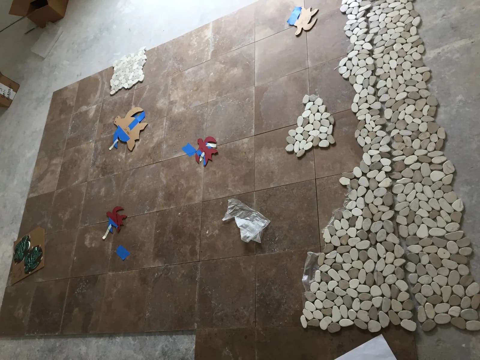 Travertine floor tile and pebble mosaic sheets dry-laid on a concrete slab with cardboard templates for tile accents.