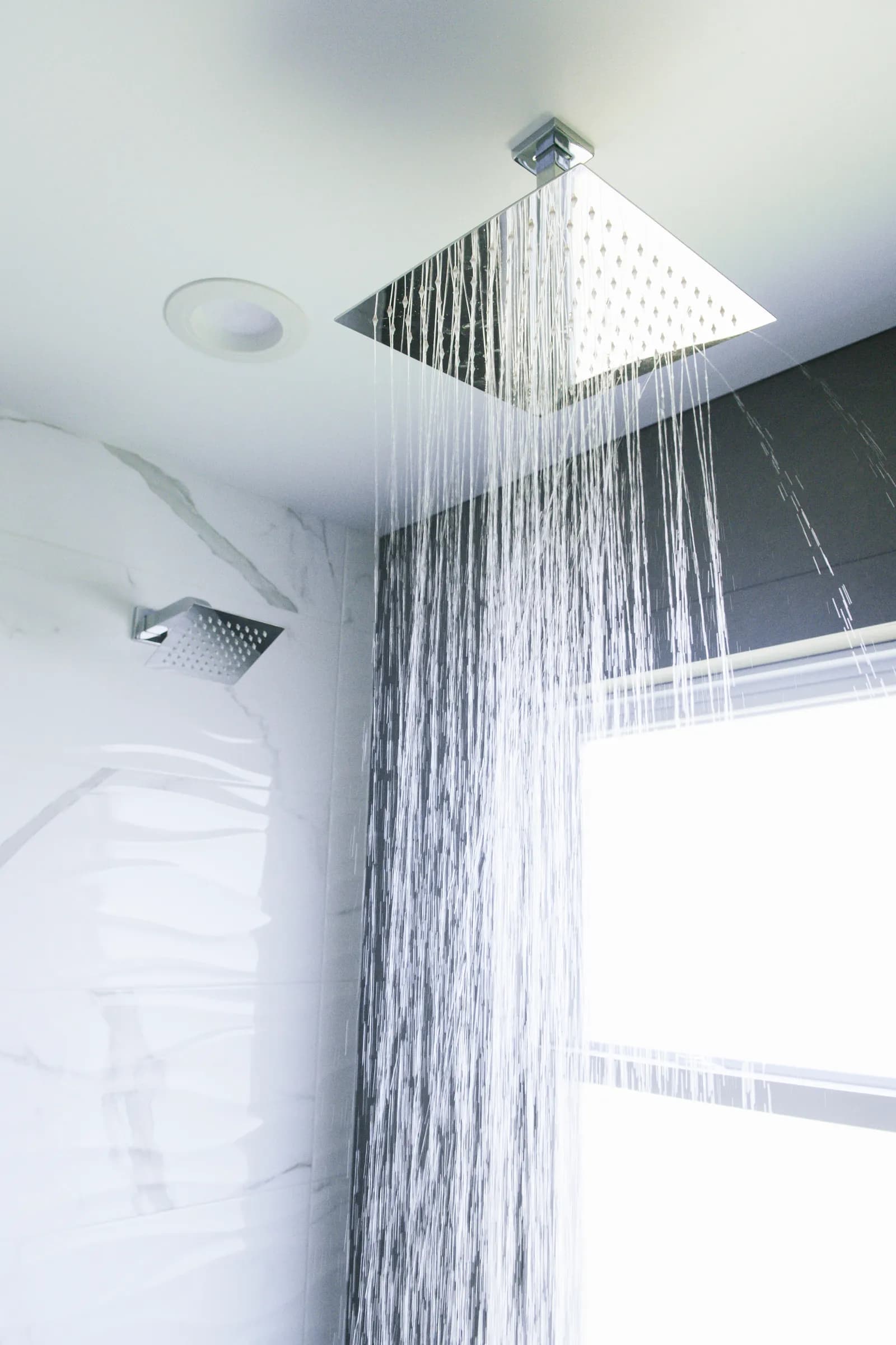Water streaming from a square ceiling-mounted rain shower head beside a wall-mounted head against marble-look tile.