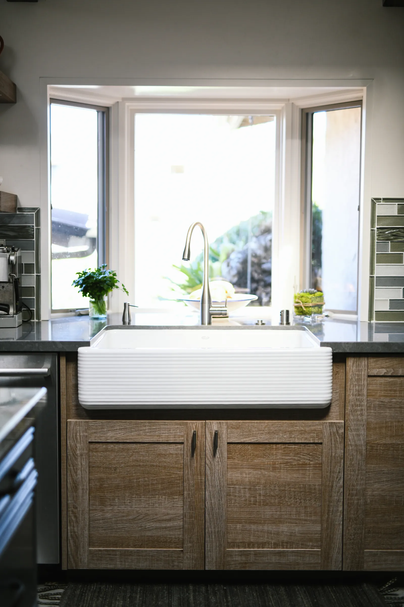 Close-up of a white fluted farmhouse sink with a gooseneck faucet below a bay window.