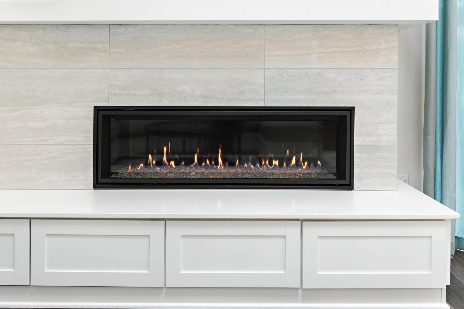Linear gas fireplace set in a light stone tile wall above white built-in cabinetry.