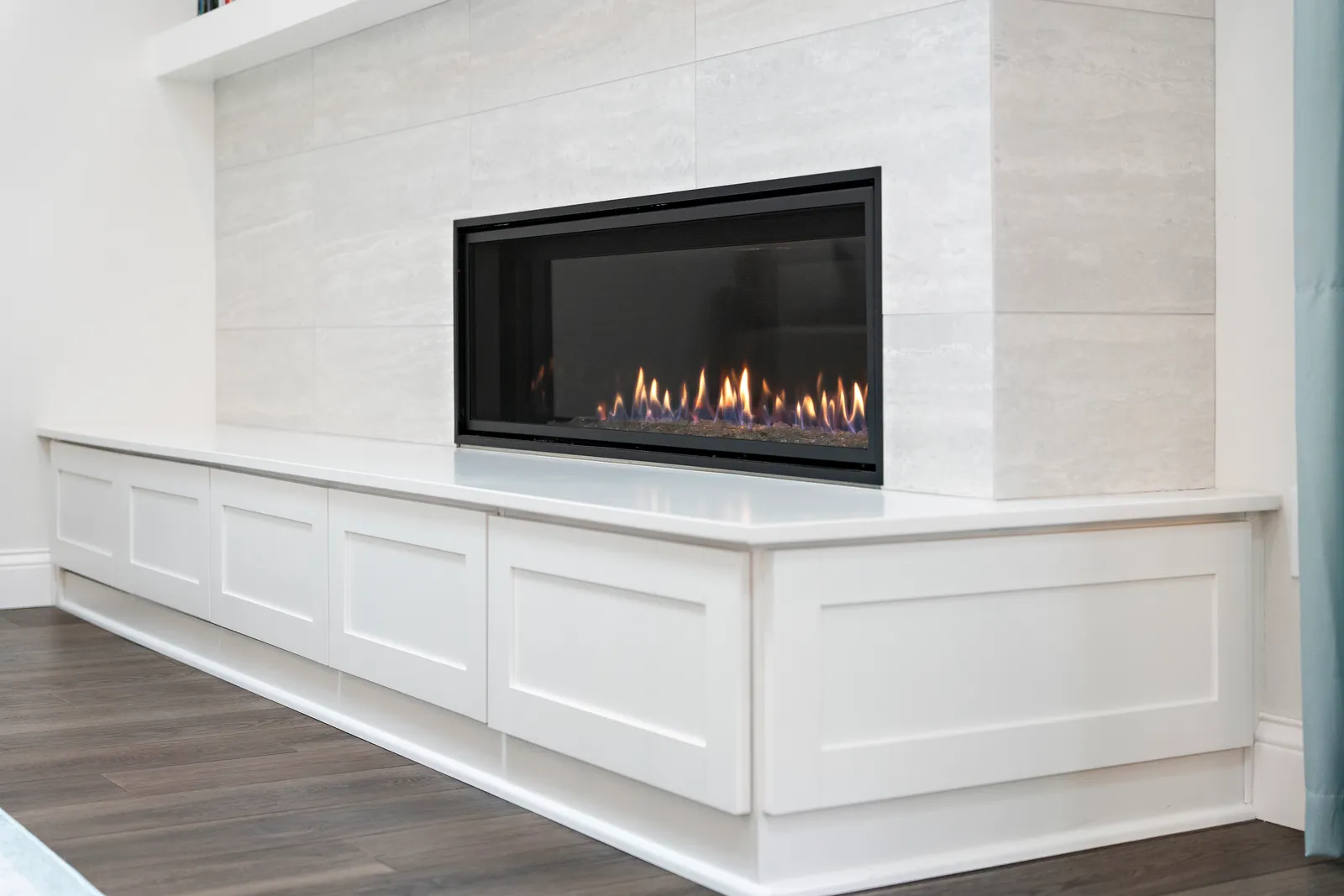 A linear gas fireplace set in light stone-look tile above a long white cabinet bench.