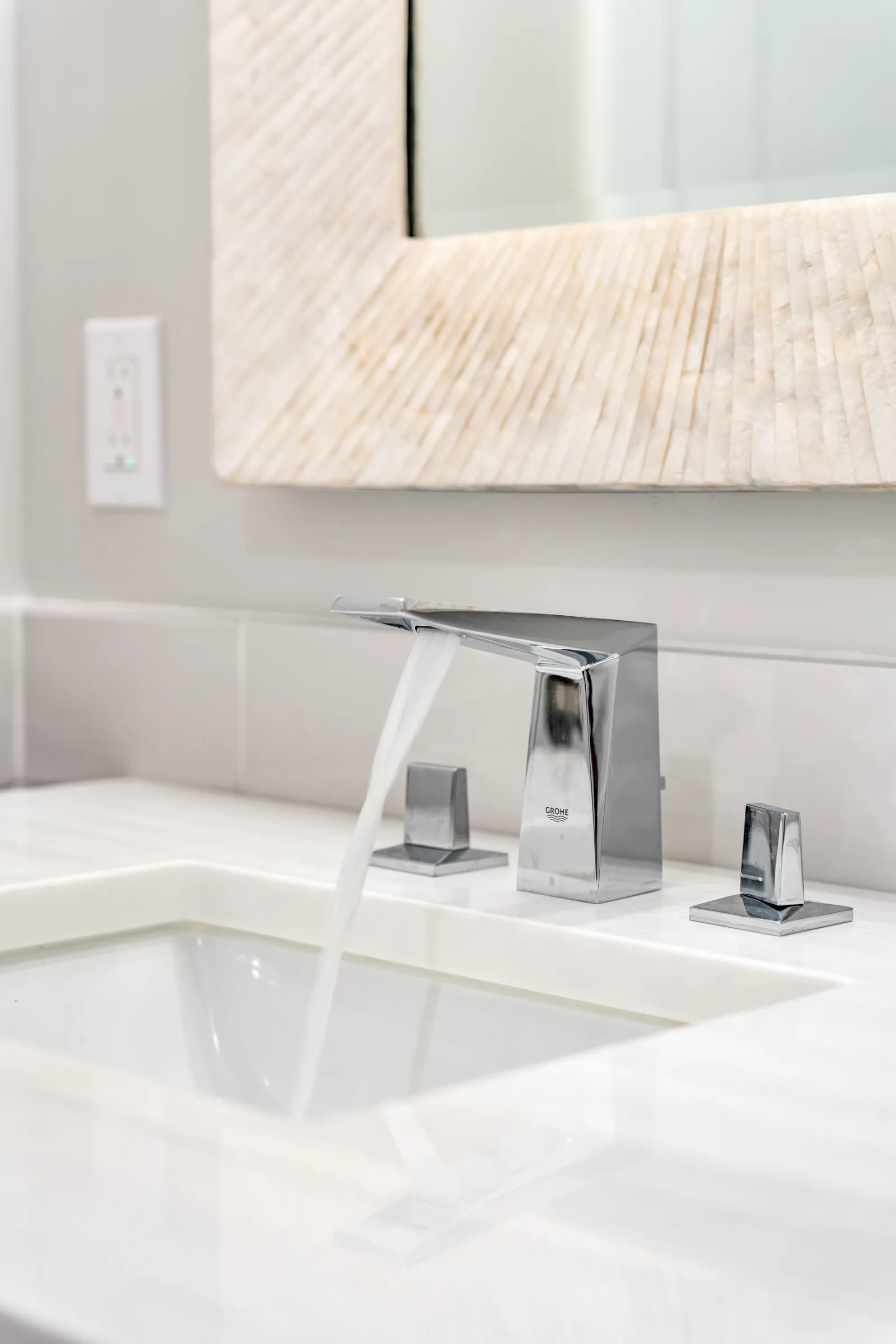 Chrome widespread faucet running water into an undermount sink below a stone-framed mirror.