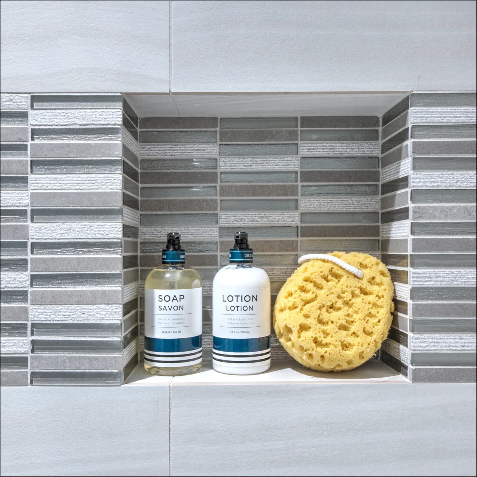 Shower niche lined with gray glass and stone mosaic tile, styled with soap and lotion pump bottles and a yellow sea sponge.