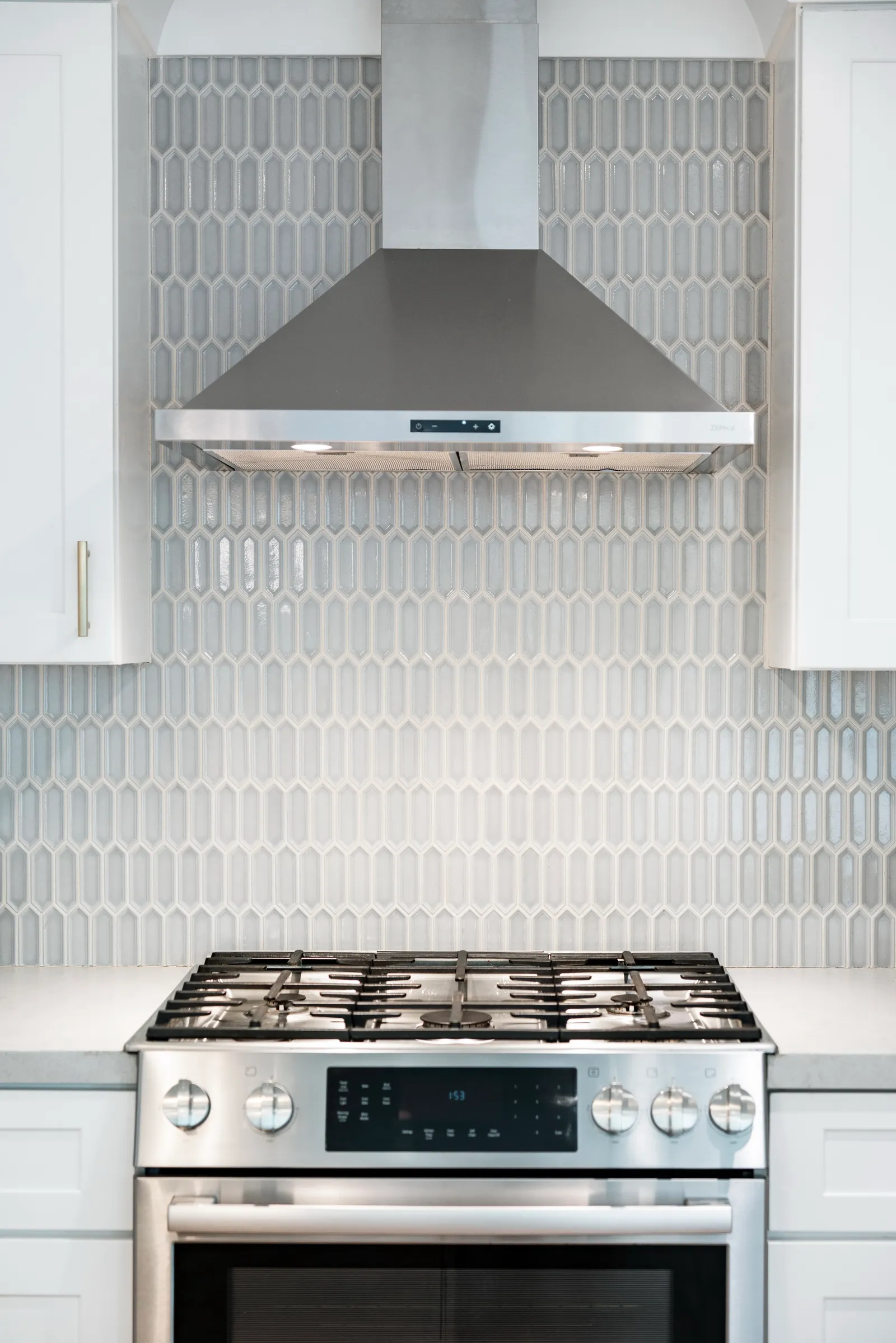 Stainless range hood and gas range against a gray picket-shaped tile backsplash flanked by white shaker cabinets.