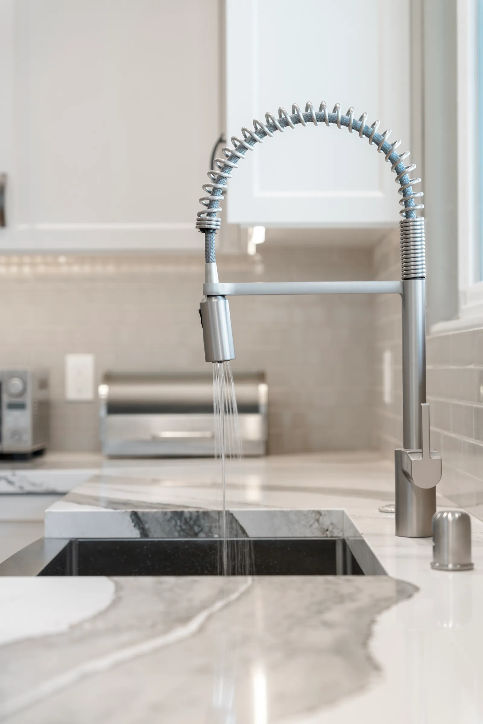 Close-up of a stainless spring-coil kitchen faucet running water over an undermount sink in a marbled quartz counter.