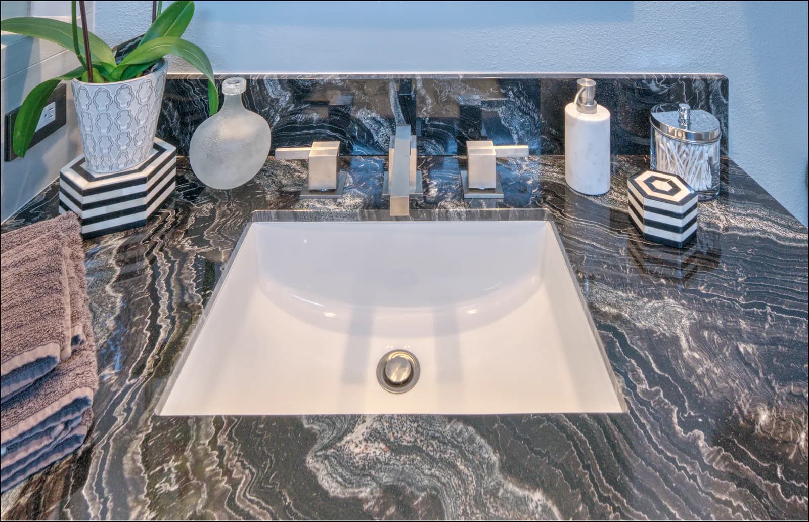 A close-up of a white undermount sink in a dramatic black-and-white veined granite counter with square modern faucet handles.