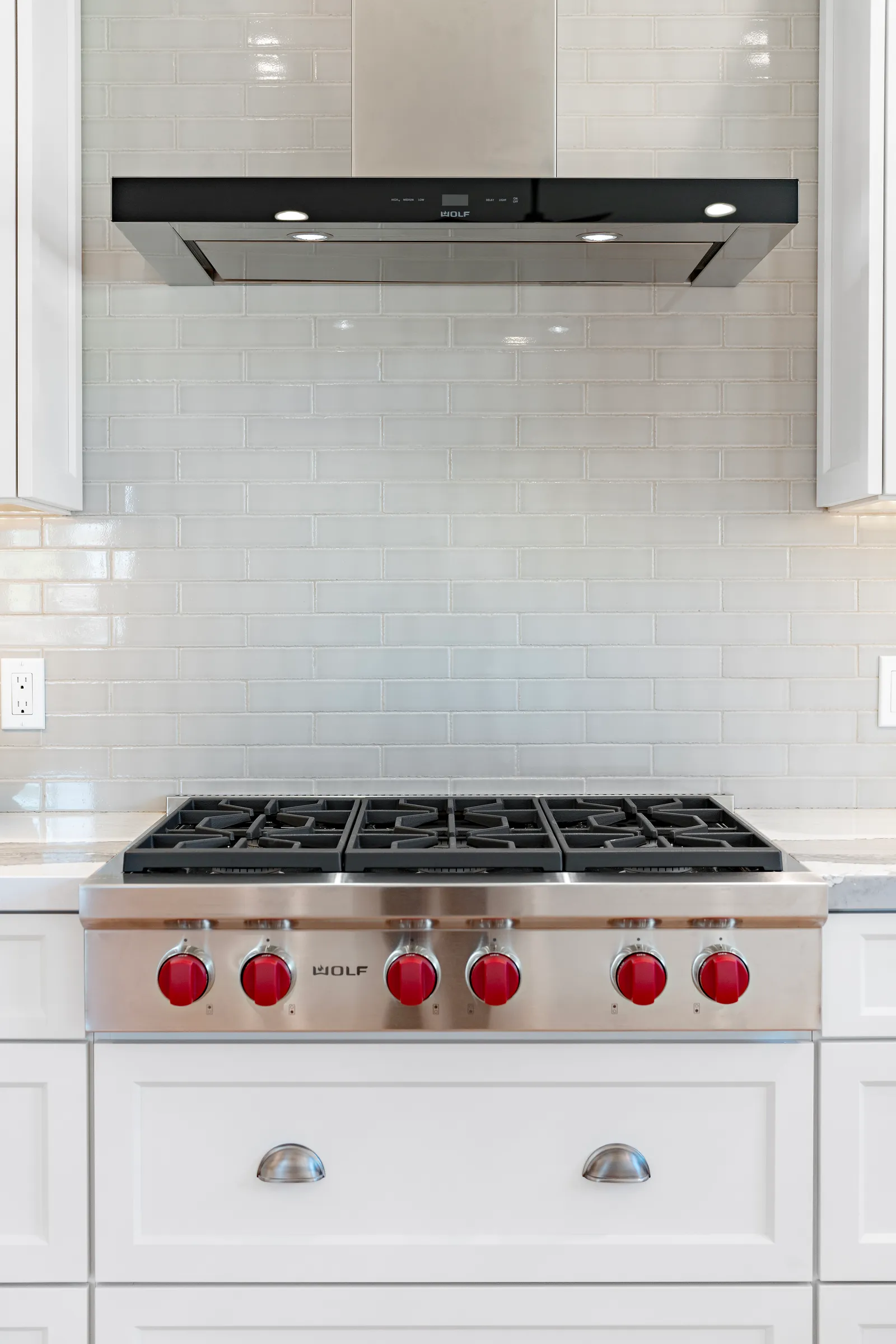 Stainless steel gas rangetop with red knobs below a vent hood and white subway tile backsplash.