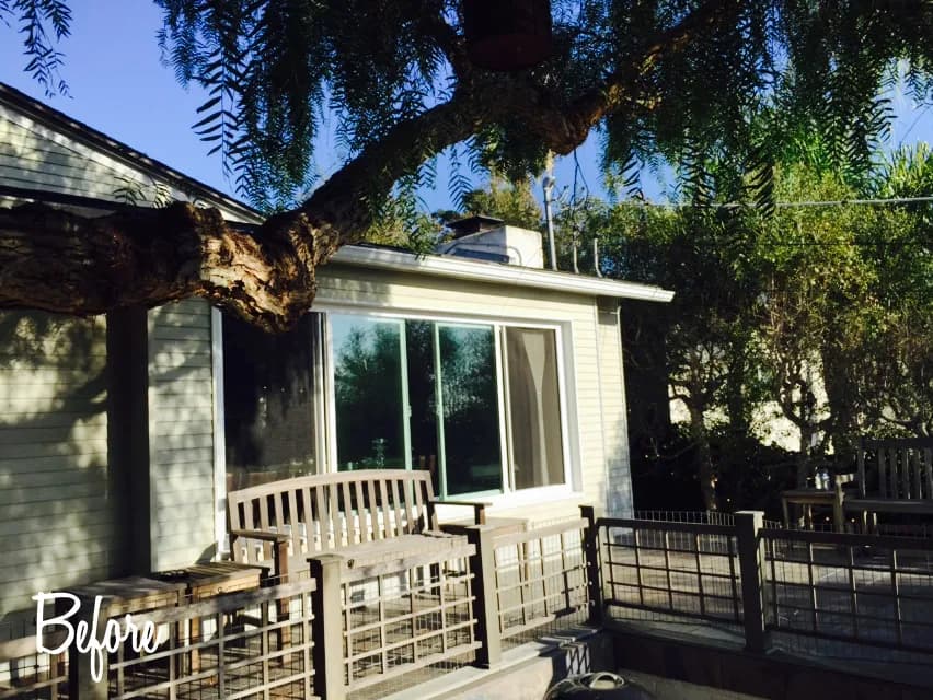 The rear of an older single-story house with a large sliding window, wood deck railing and an overhanging tree, with a Before caption overlaid.