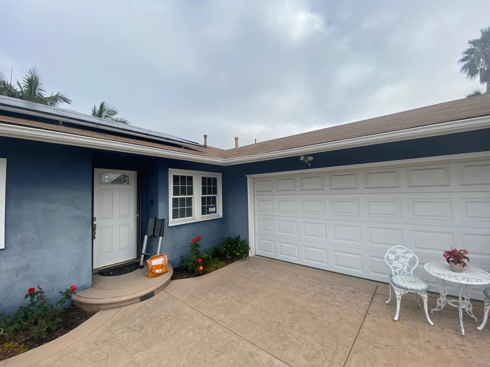 A single-story blue stucco house with a white garage door, rose bushes and a stamped concrete driveway and entry step under a cloudy sky.