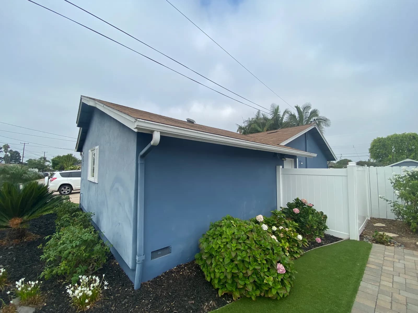 The side of a blue stucco house with a brown shingle roof, white vinyl fence, and landscaped beds.