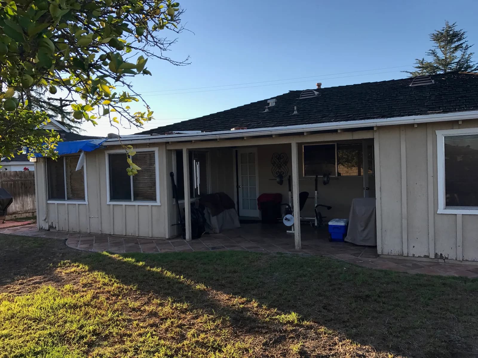 Rear of an older single-story house with a covered patio, worn siding, a tarped roof section, and patchy lawn at dusk.