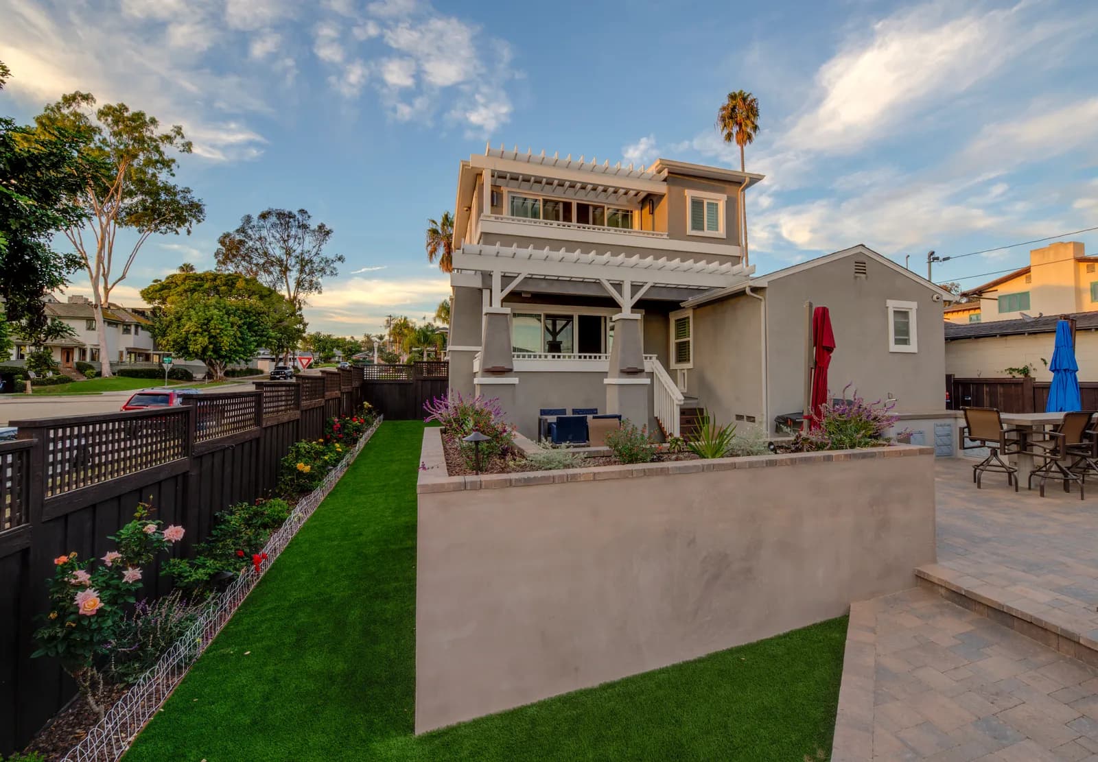 Two-story gray craftsman-style home at dusk with white pergola details, artificial turf side yard, and rose garden along a dark wood fence.