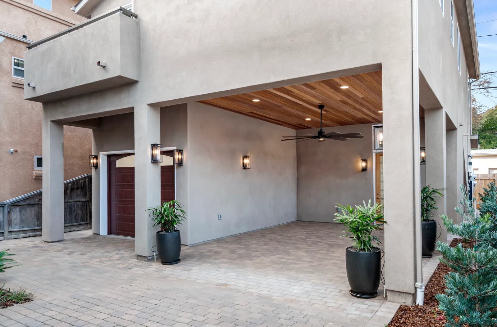 A modern stucco home exterior with an open carport, wood plank ceiling, paver driveway, and potted plants.