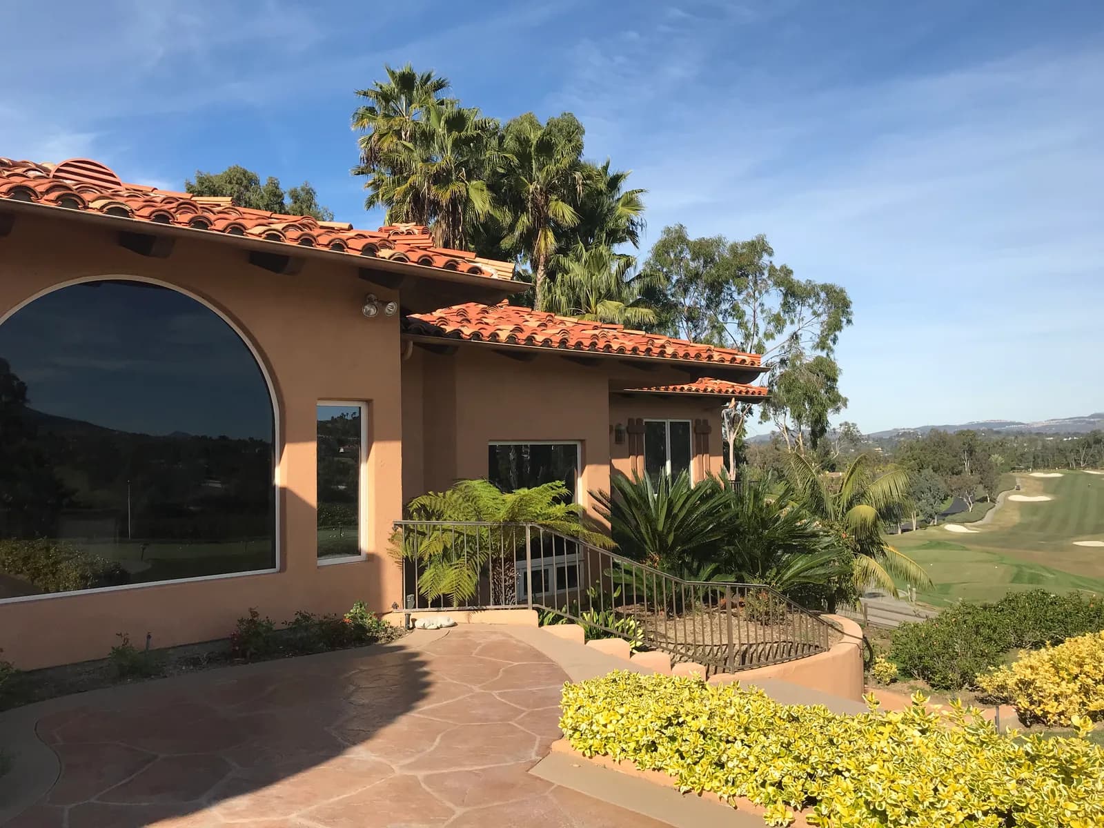 Spanish-style home with large arched window, clay tile roof, palm trees, and a golf course view beyond.