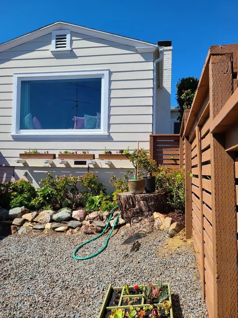 White horizontal-siding cottage with a large picture window, planter boxes, and a gravel side yard.