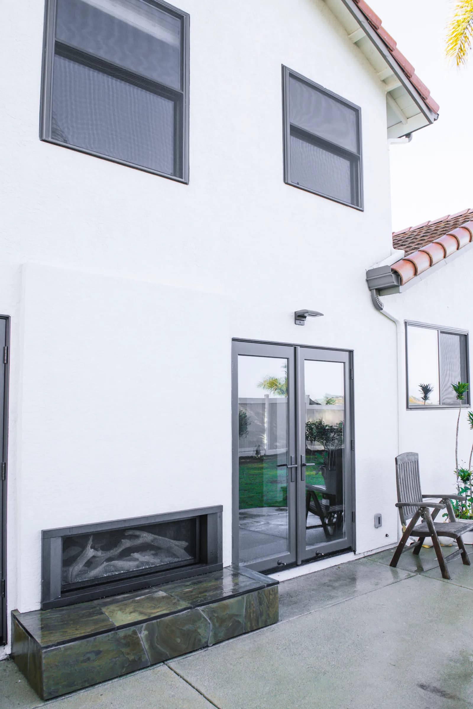 The rear of a white stucco two-story home with dark-framed windows, French patio doors, a linear outdoor fireplace with slate hearth and a wood patio chair.