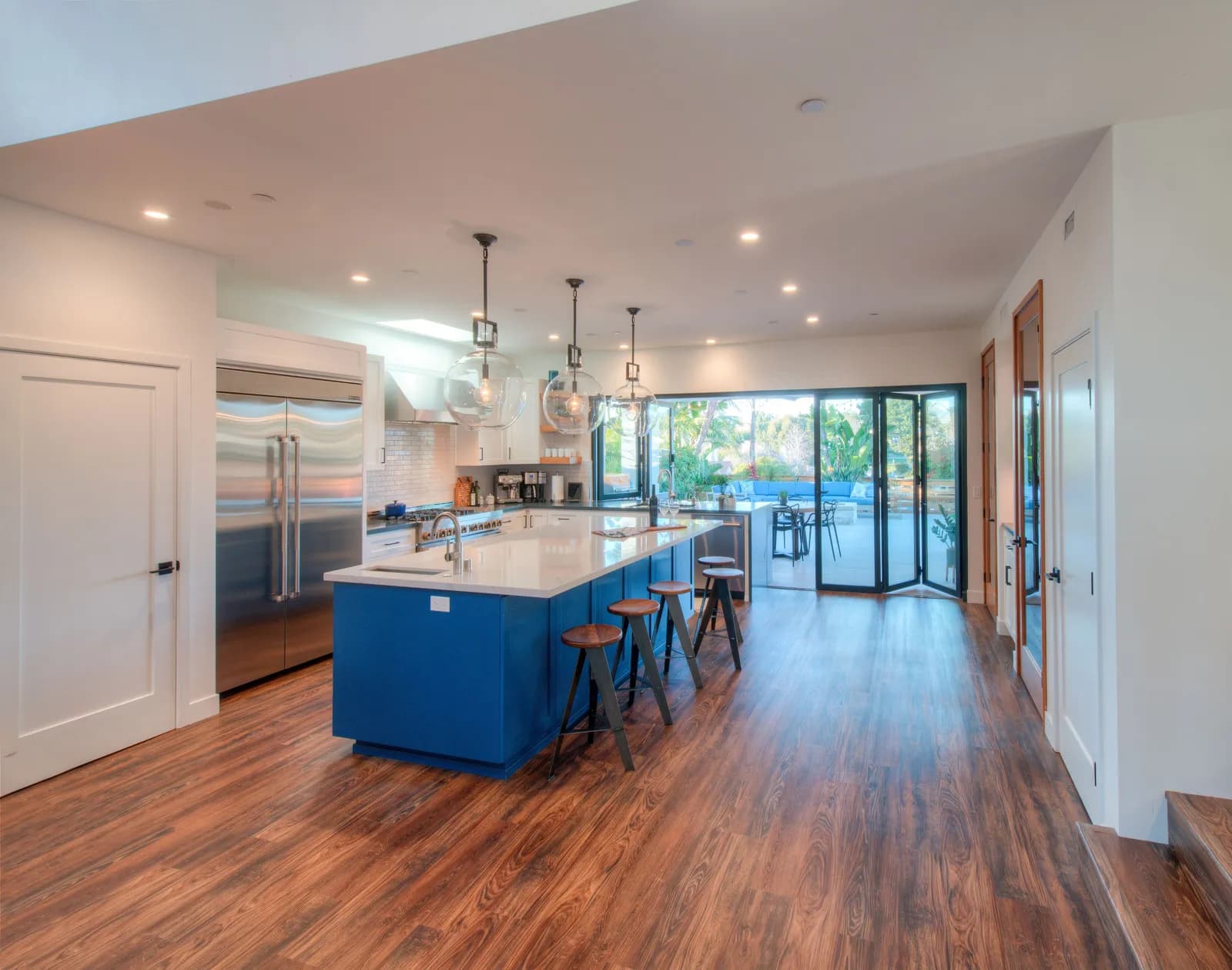 A kitchen with a long blue island, glass globe pendants, stainless refrigerator, and folding glass doors opening to a patio.