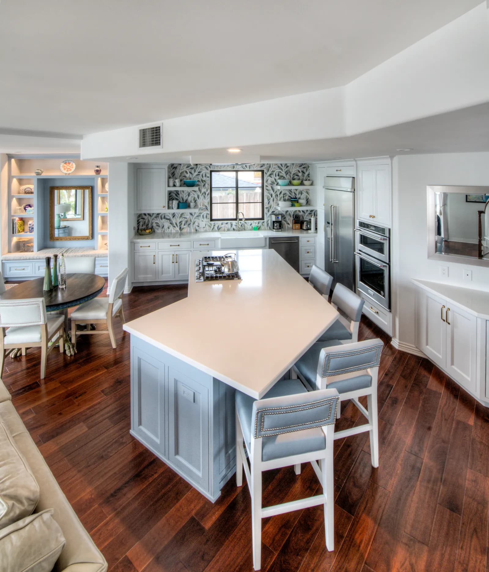 An open-plan kitchen with a pale blue island, white countertops, patterned tile backsplash, and upholstered bar stools.