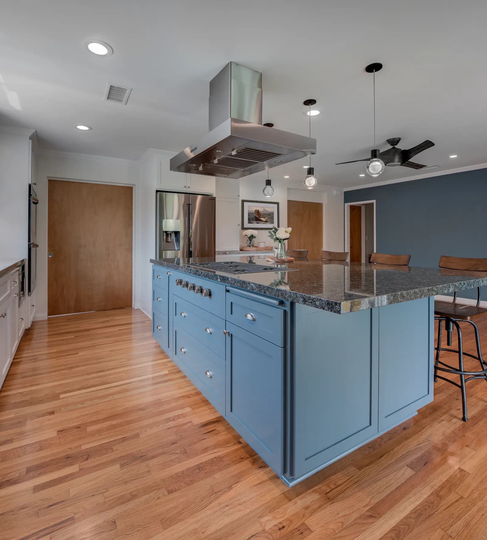 A slate blue kitchen island with granite counter, built-in cooktop, and a stainless island hood over light oak floors.