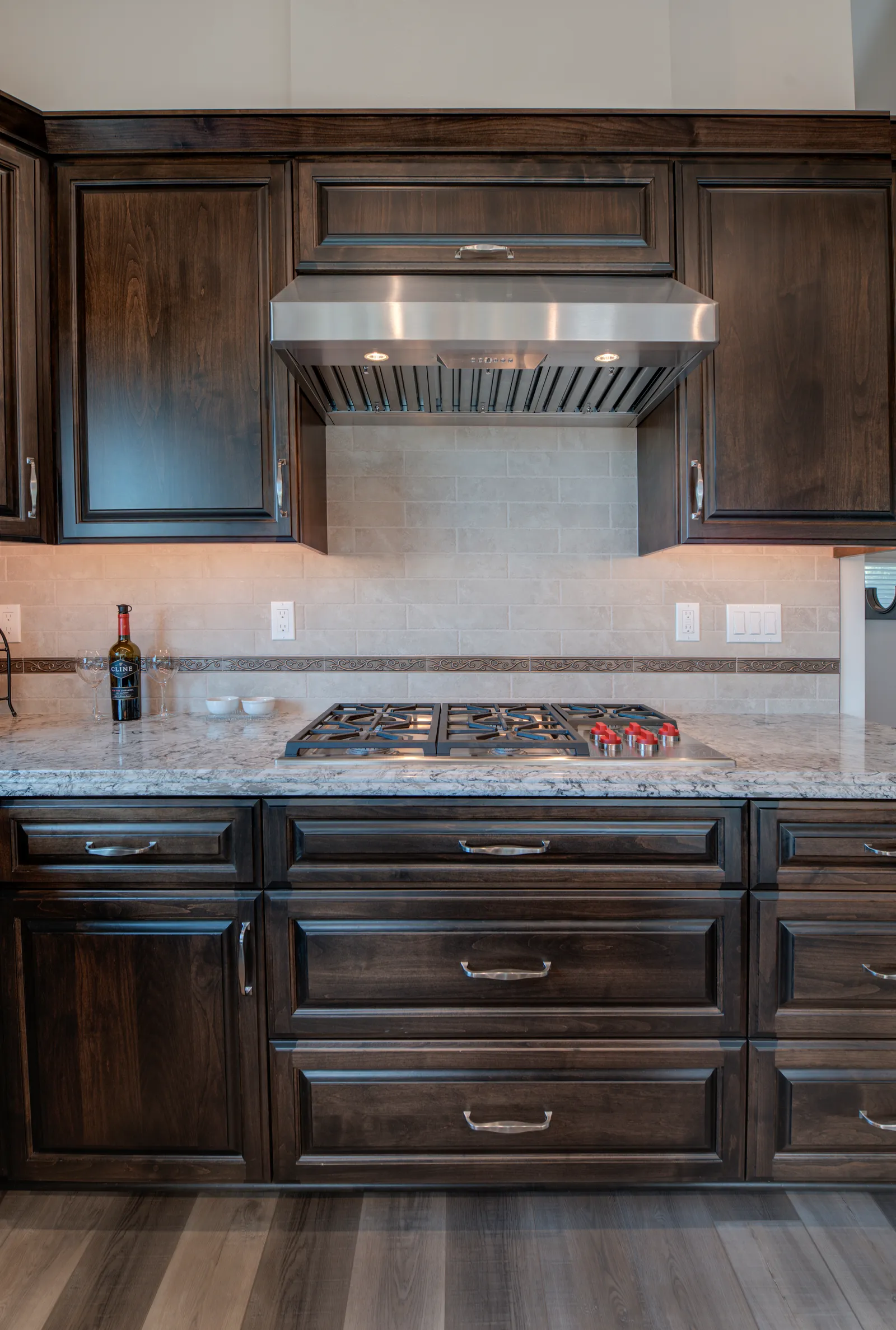 Dark stained cabinetry surrounds a stainless range hood and gas cooktop set in a granite counter.