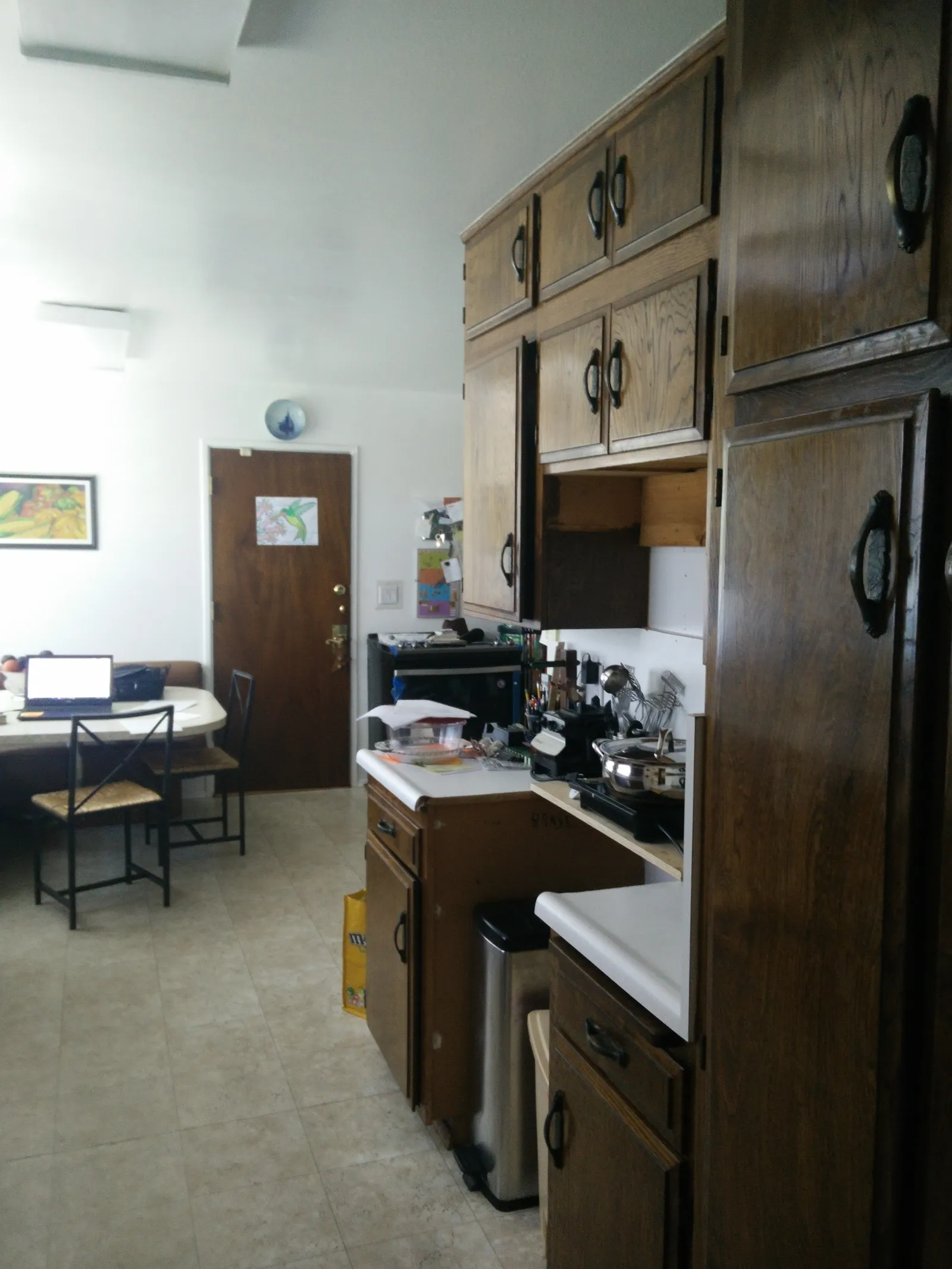 Galley kitchen with dark-stained oak cabinets, white laminate counters, and an adjacent dining table before renovation.