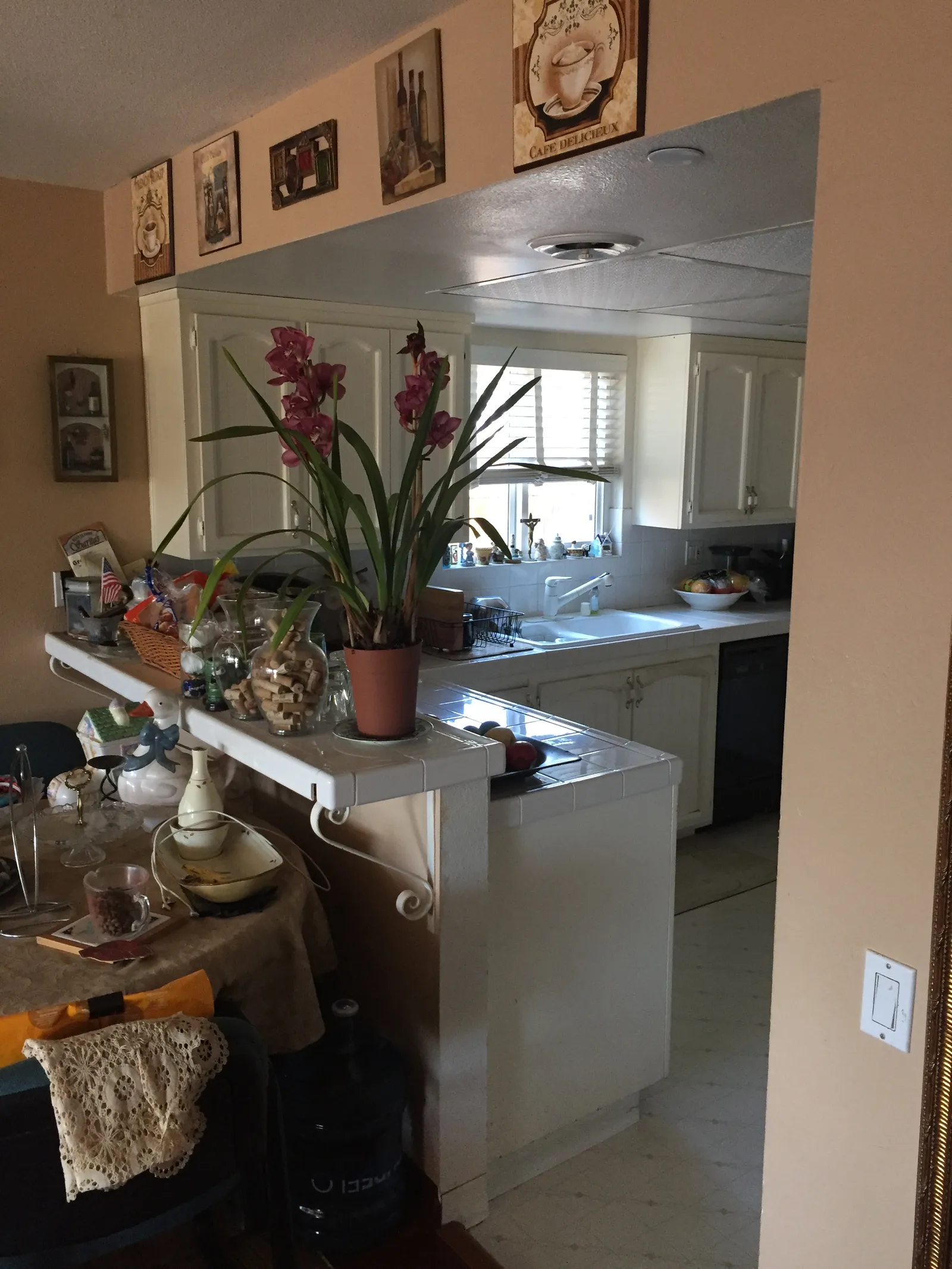 Dated kitchen seen past a tile pass-through counter with white cabinets, tile countertops, and cluttered shelves.
