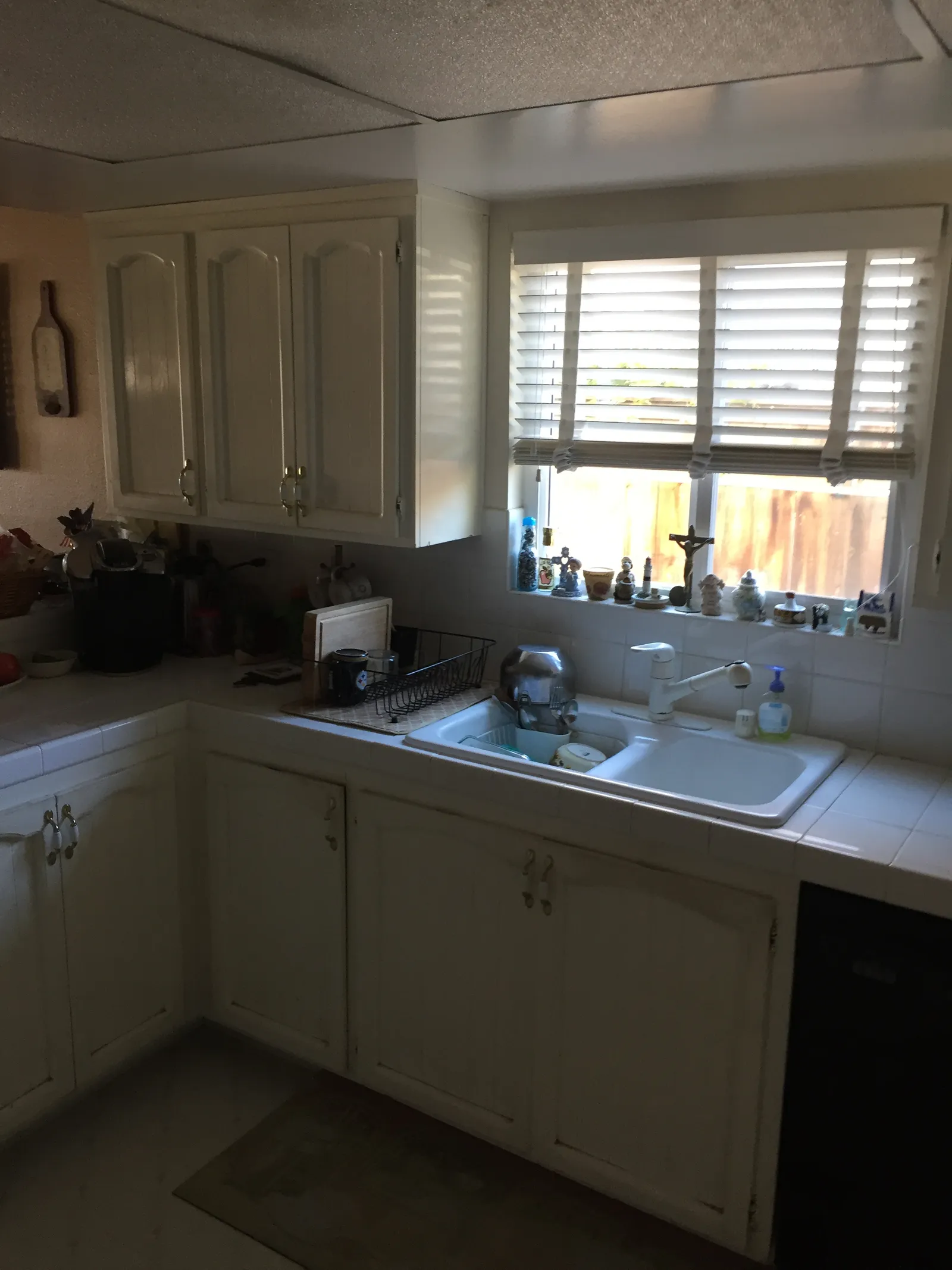 A dated kitchen with cream raised-panel cabinets, white tile countertops, and a double sink below a window with blinds.