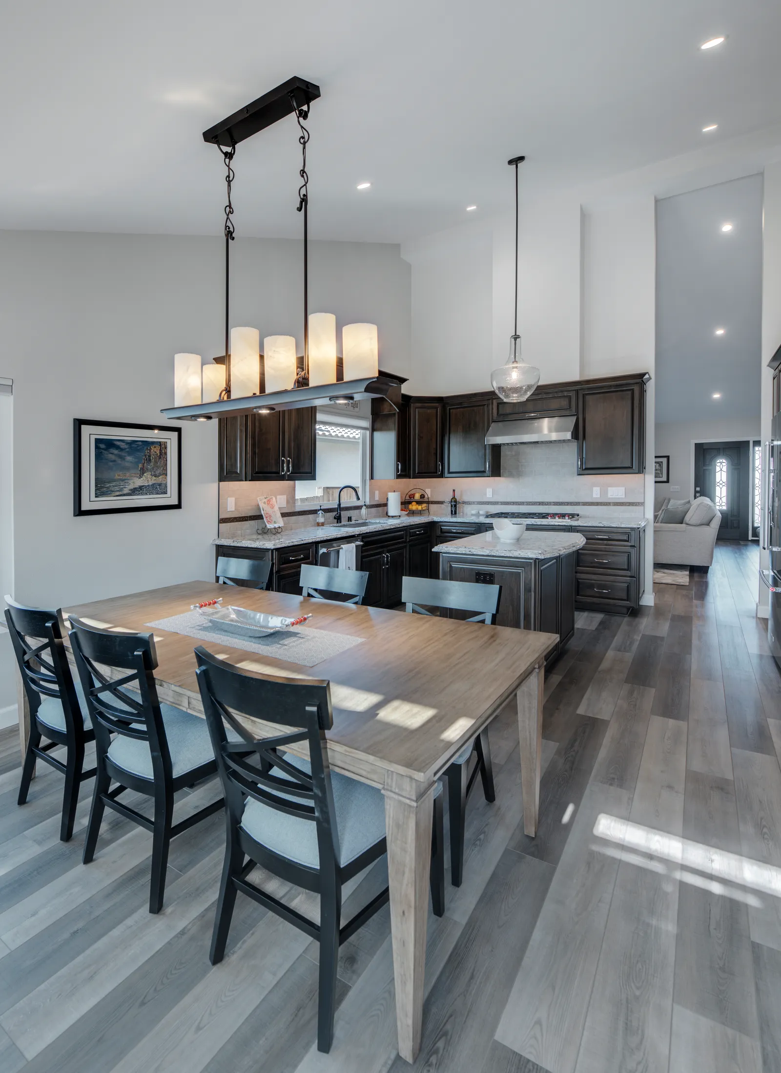 A dining table with black cross-back chairs under a candle-style chandelier, with a dark-cabinet kitchen and gray wood-look floors beyond.