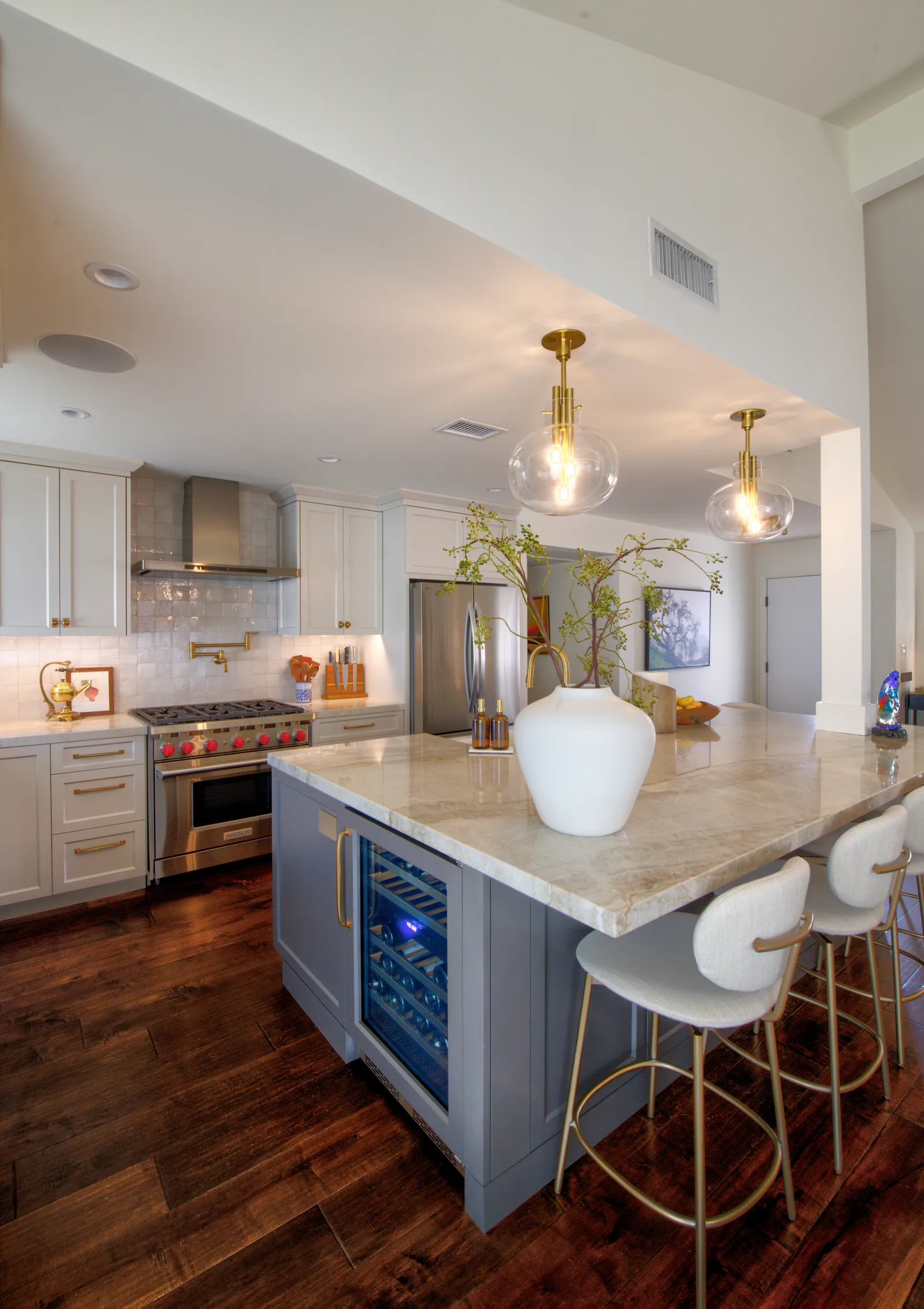 A kitchen with a gray island housing a glass-door wine refrigerator, brass globe pendants, and a red-knob professional range.