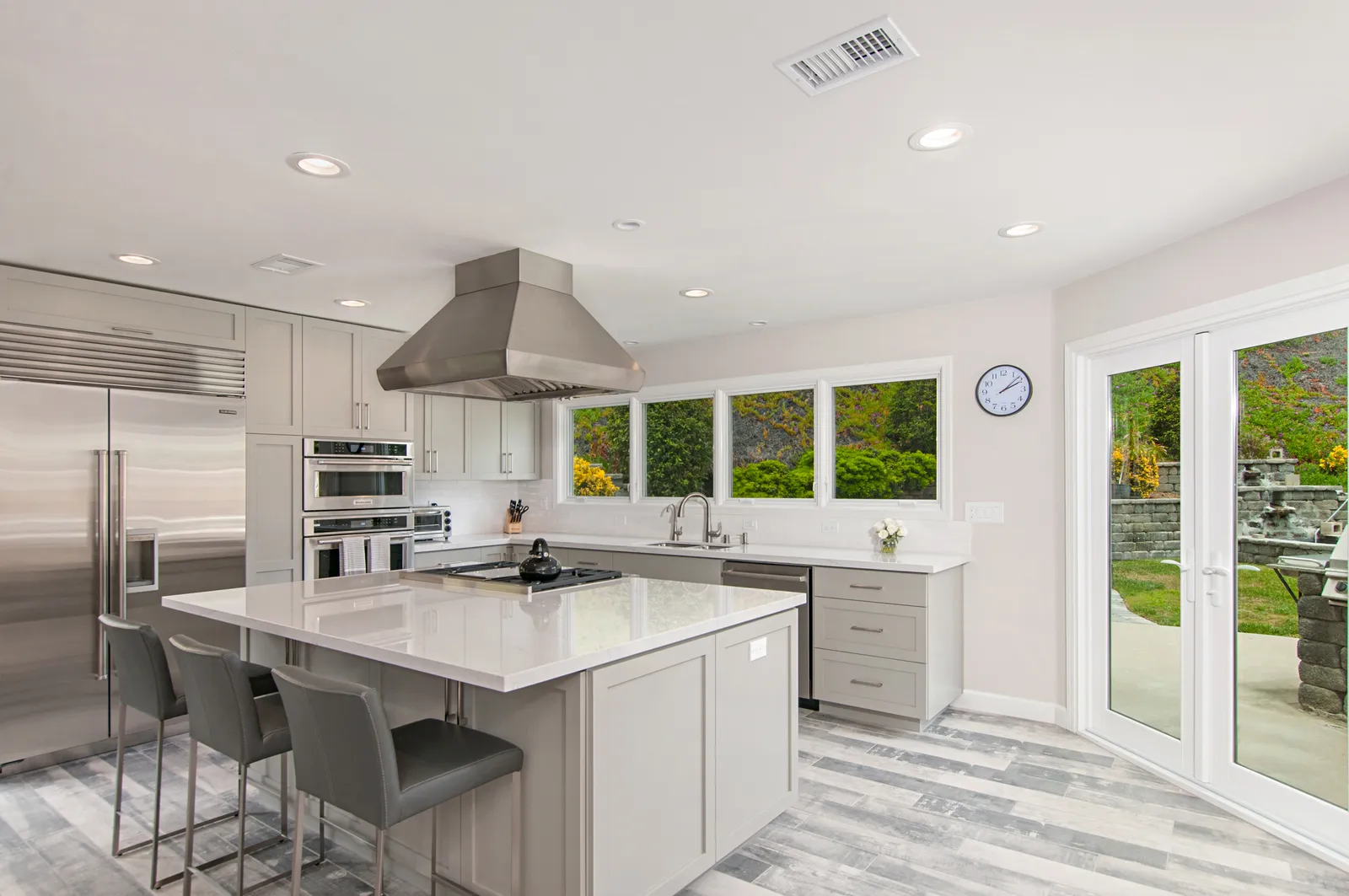A light gray kitchen with a large island, stainless chimney hood, French doors to a patio, and gray wood-look tile floors.
