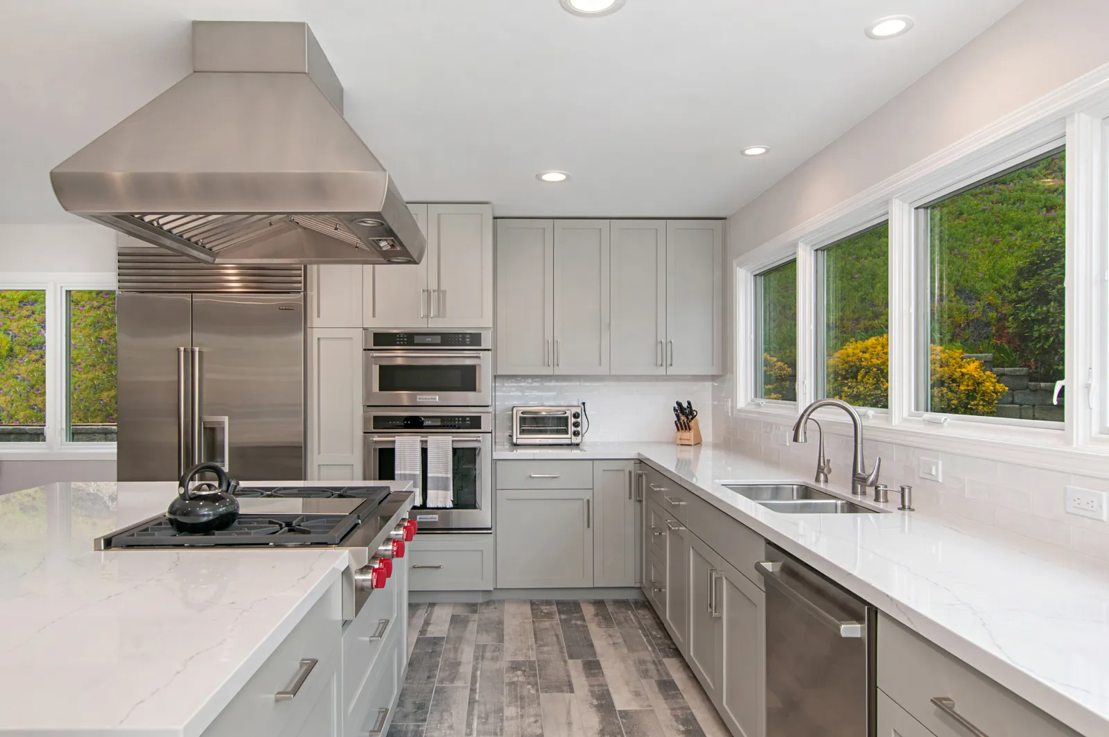 A remodeled kitchen with light gray cabinets, white quartz counters, a stainless hood over a pro range, and wide windows over the sink.