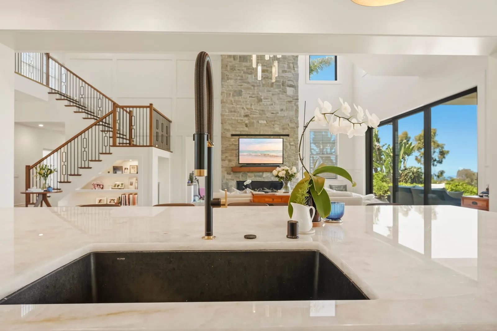 A kitchen island sink with a black spring faucet looking out to a two-story great room with a stone fireplace and staircase.