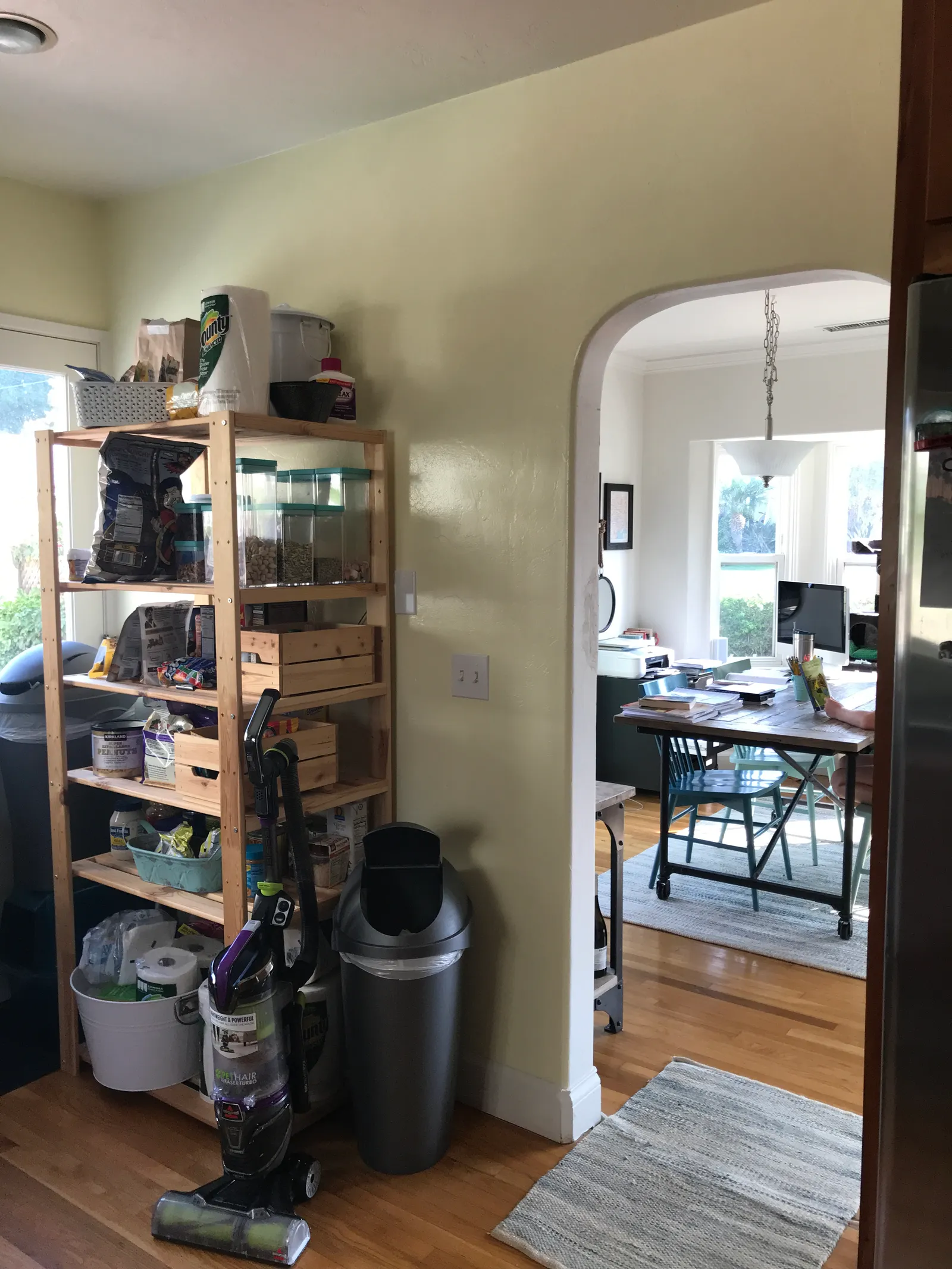 Kitchen corner with open wooden pantry shelving and an arched doorway into a sunny dining room.