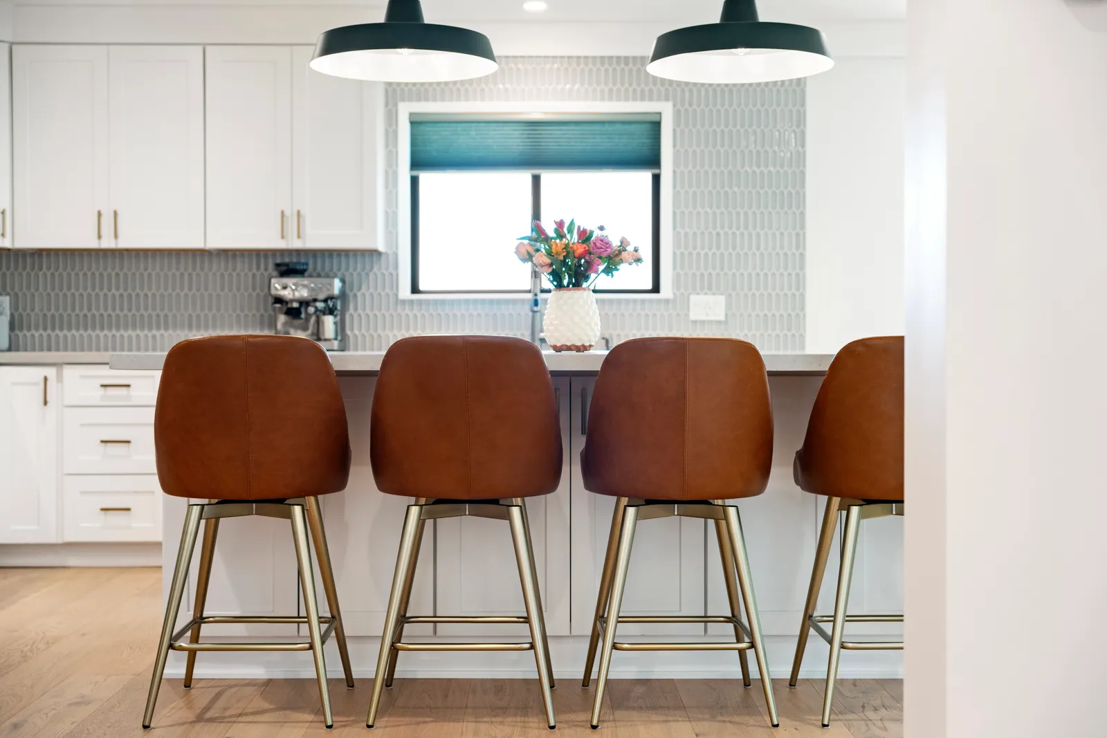 Four cognac leather counter stools with brass legs lined up at a white kitchen island under dark cone pendants.