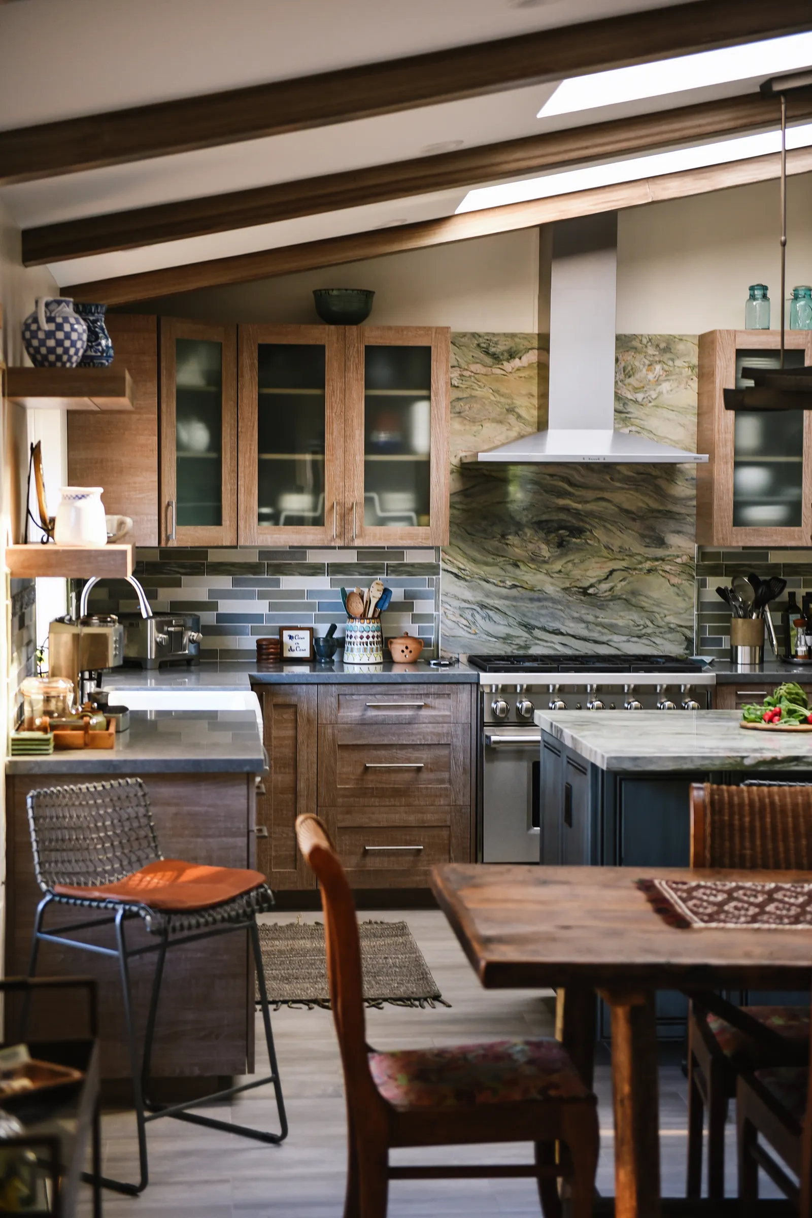 Kitchen with dramatic stone slab backsplash, stainless chimney hood and glass-front wood cabinets.