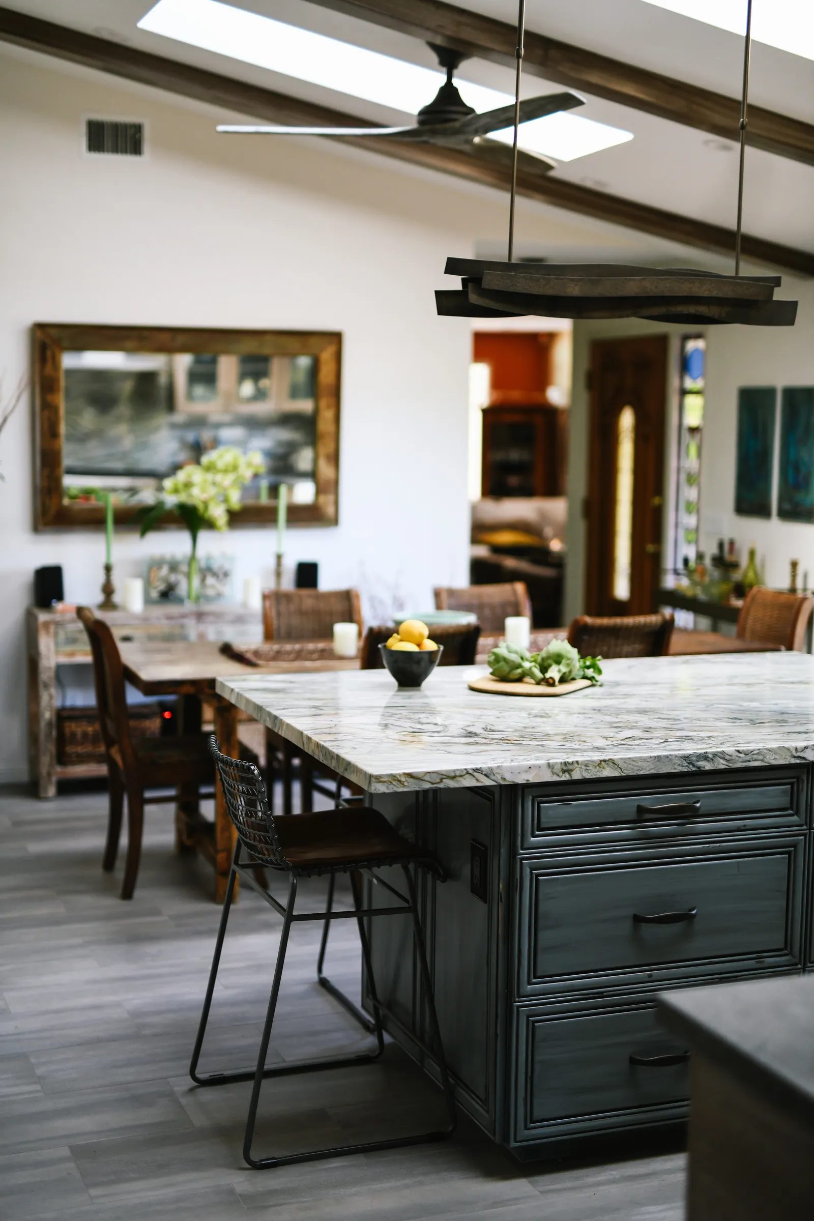 A kitchen island with a marble countertop, dark painted cabinets, and metal stools beneath skylights and wood beams.
