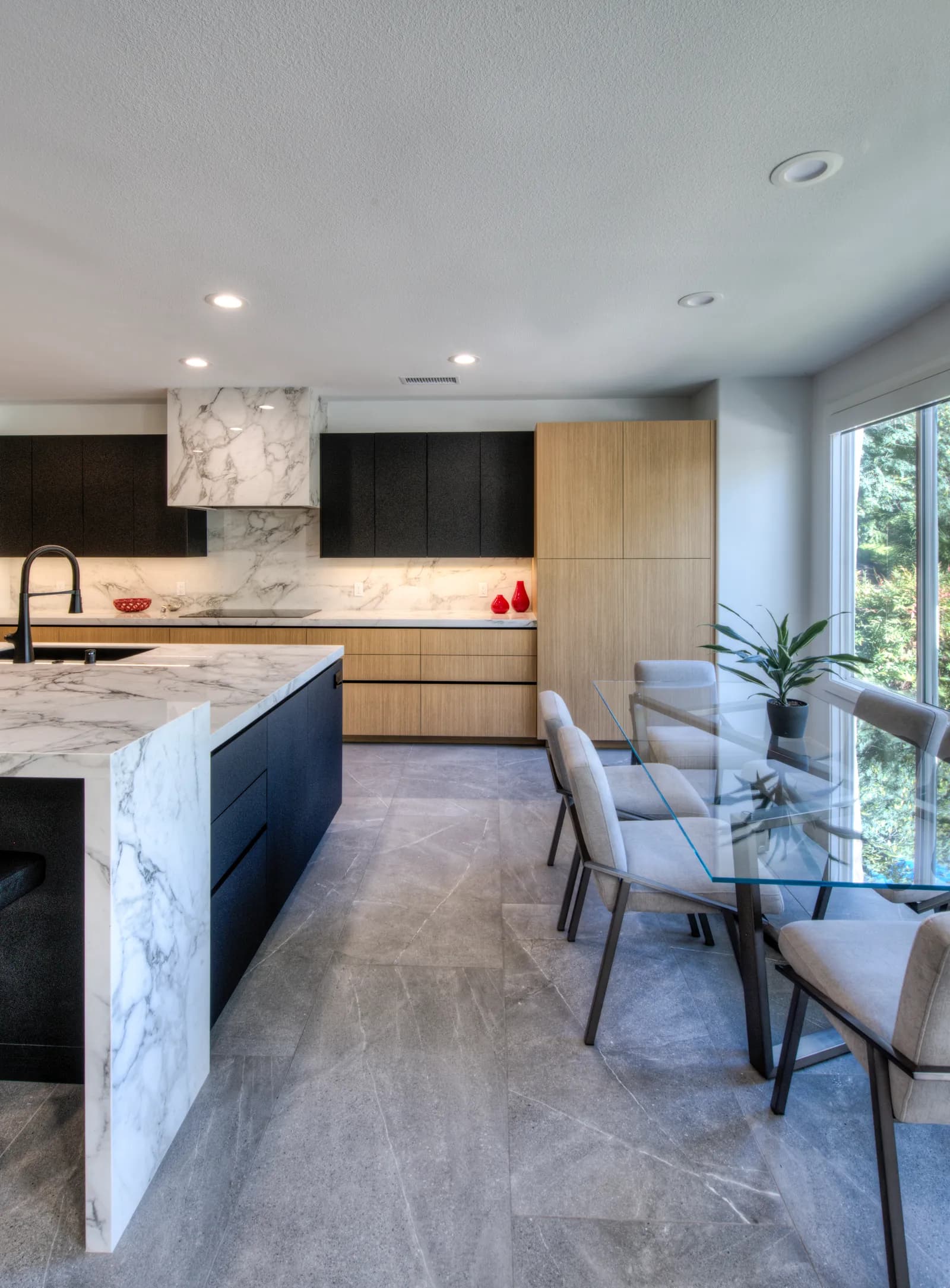 A modern kitchen with a marble waterfall island, black and oak flat-panel cabinets, and a glass dining table.