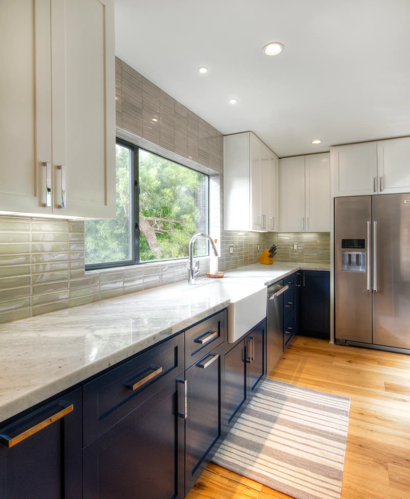 A kitchen with navy lower cabinets, white farmhouse sink, marble countertops, green glass tile backsplash, and stainless refrigerator.