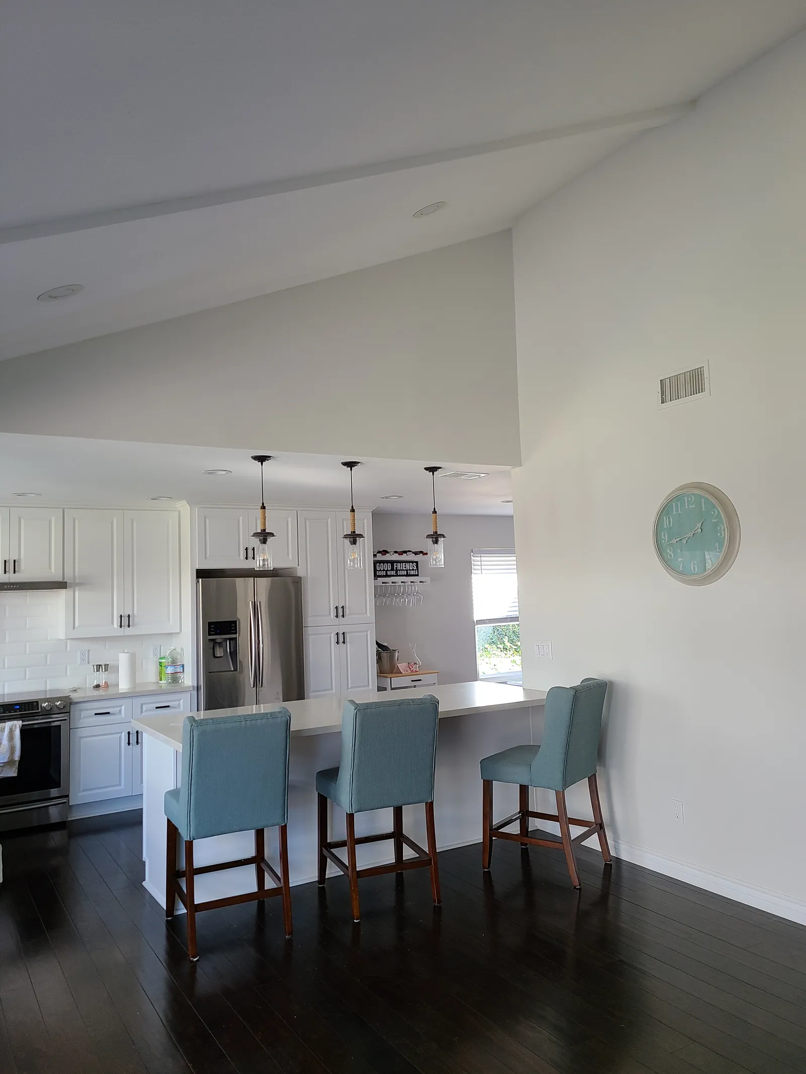 An open-plan kitchen with white raised-panel cabinets, a stainless refrigerator, pendant lights and three teal upholstered bar stools at a white counter.