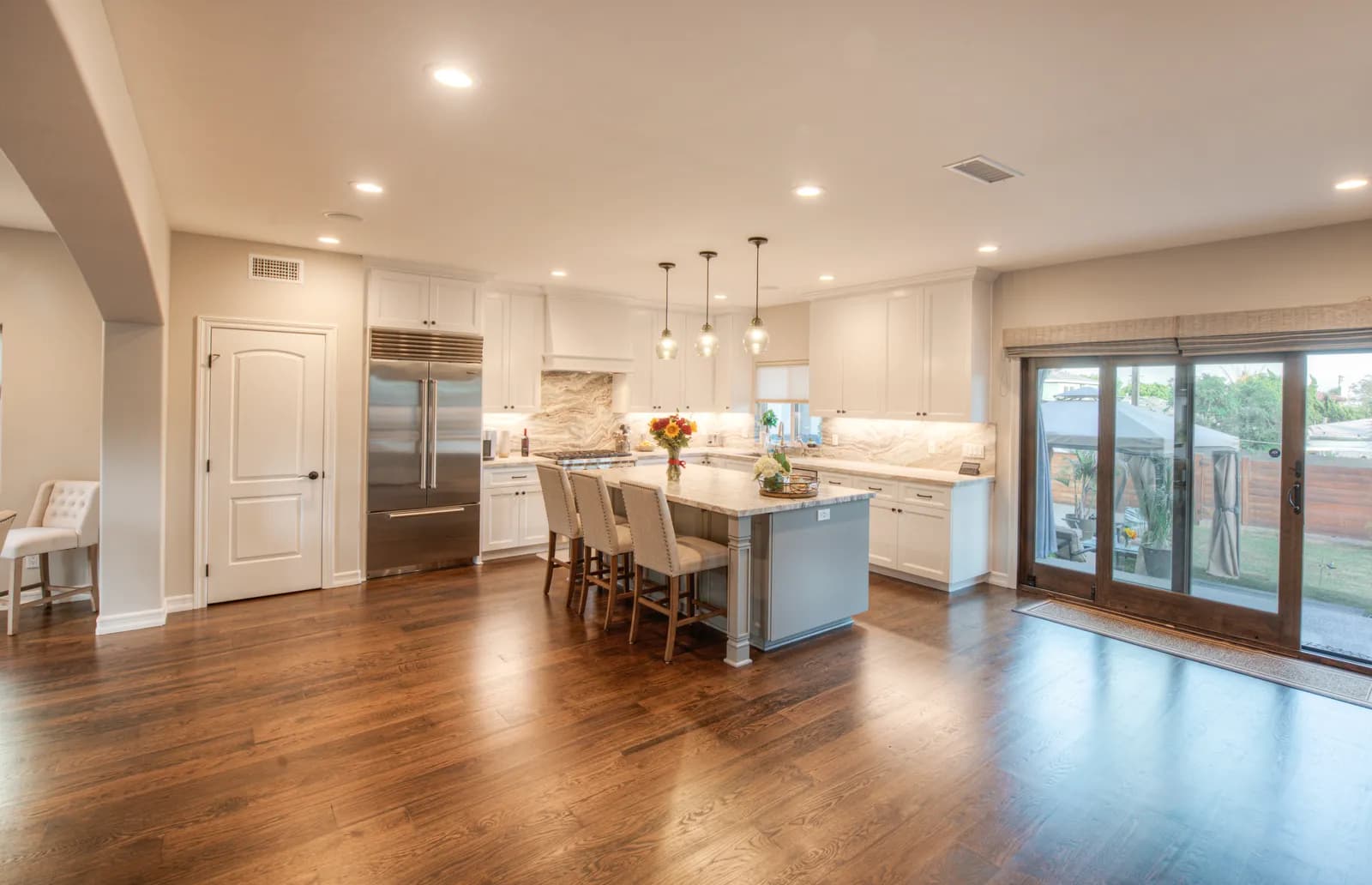 An open-plan kitchen with white cabinets, a gray island with upholstered stools, pendant lights, hardwood floors, and sliding glass doors.
