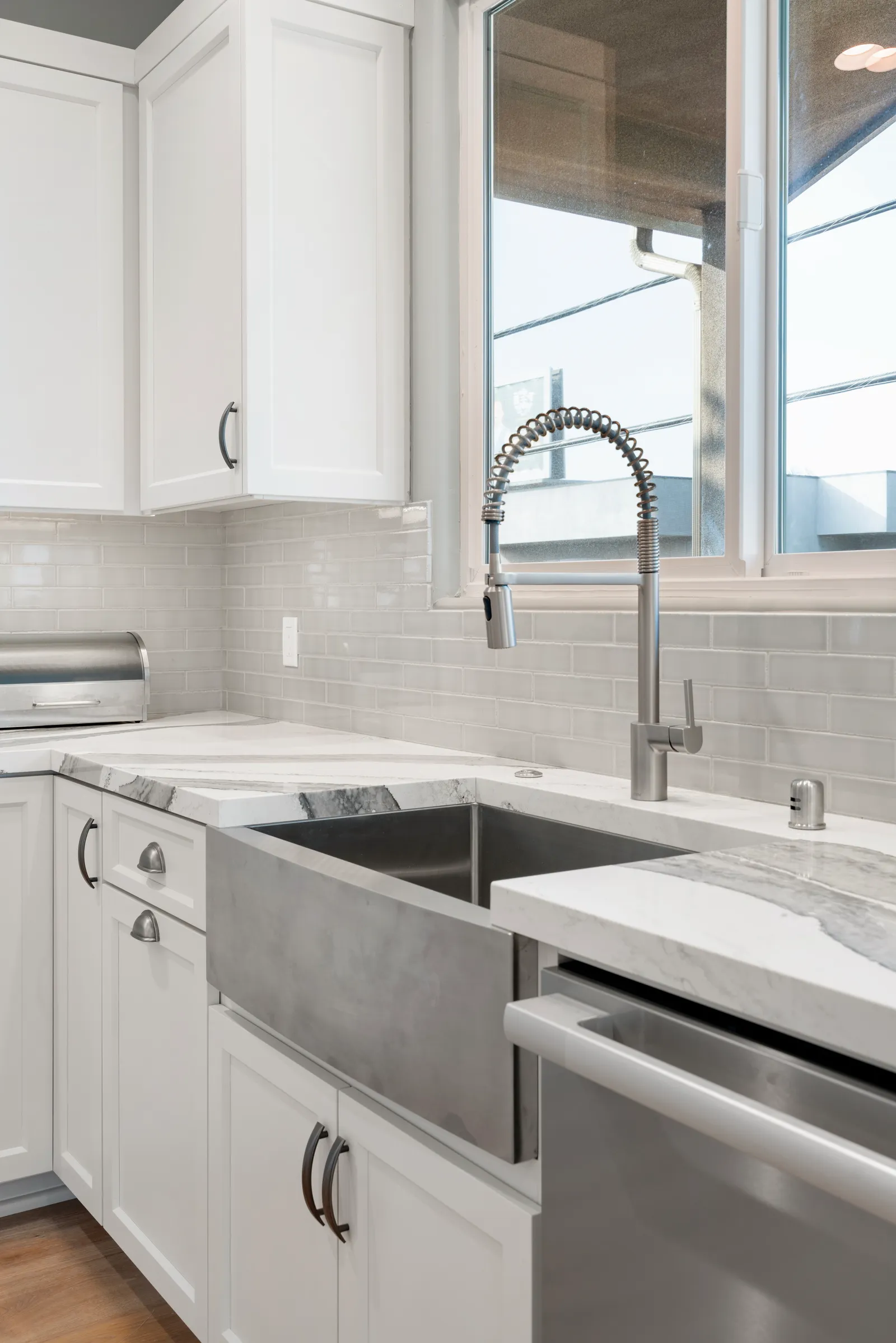 White kitchen with a stainless apron-front sink, coiled commercial-style faucet, marble-look counters, and gray subway tile.