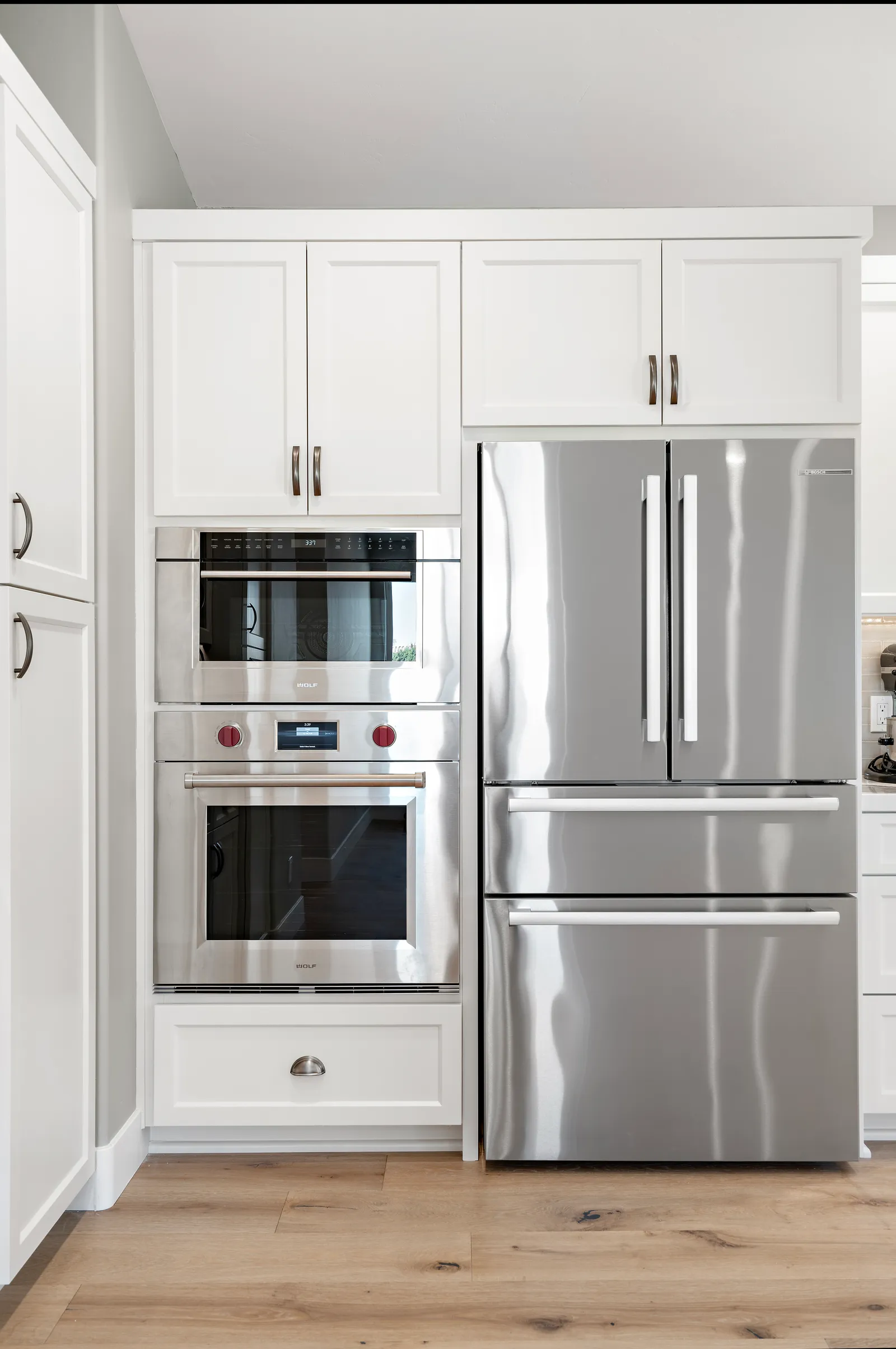 White shaker cabinetry with built-in stainless wall ovens and a French-door refrigerator over light wood flooring.