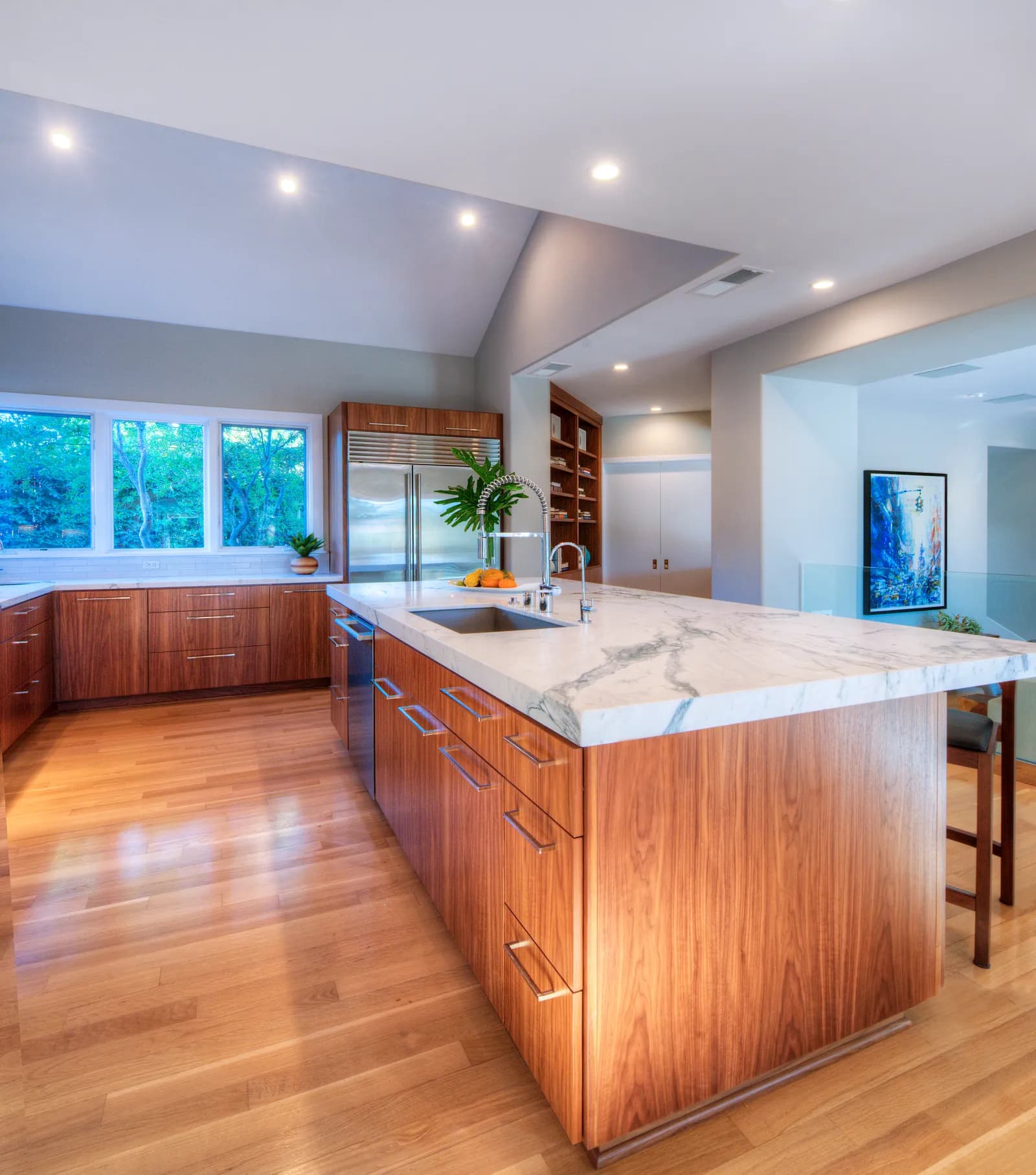 Kitchen with a long walnut island topped in veined marble, chrome spring faucet, built-in stainless refrigerator, and oak floors.