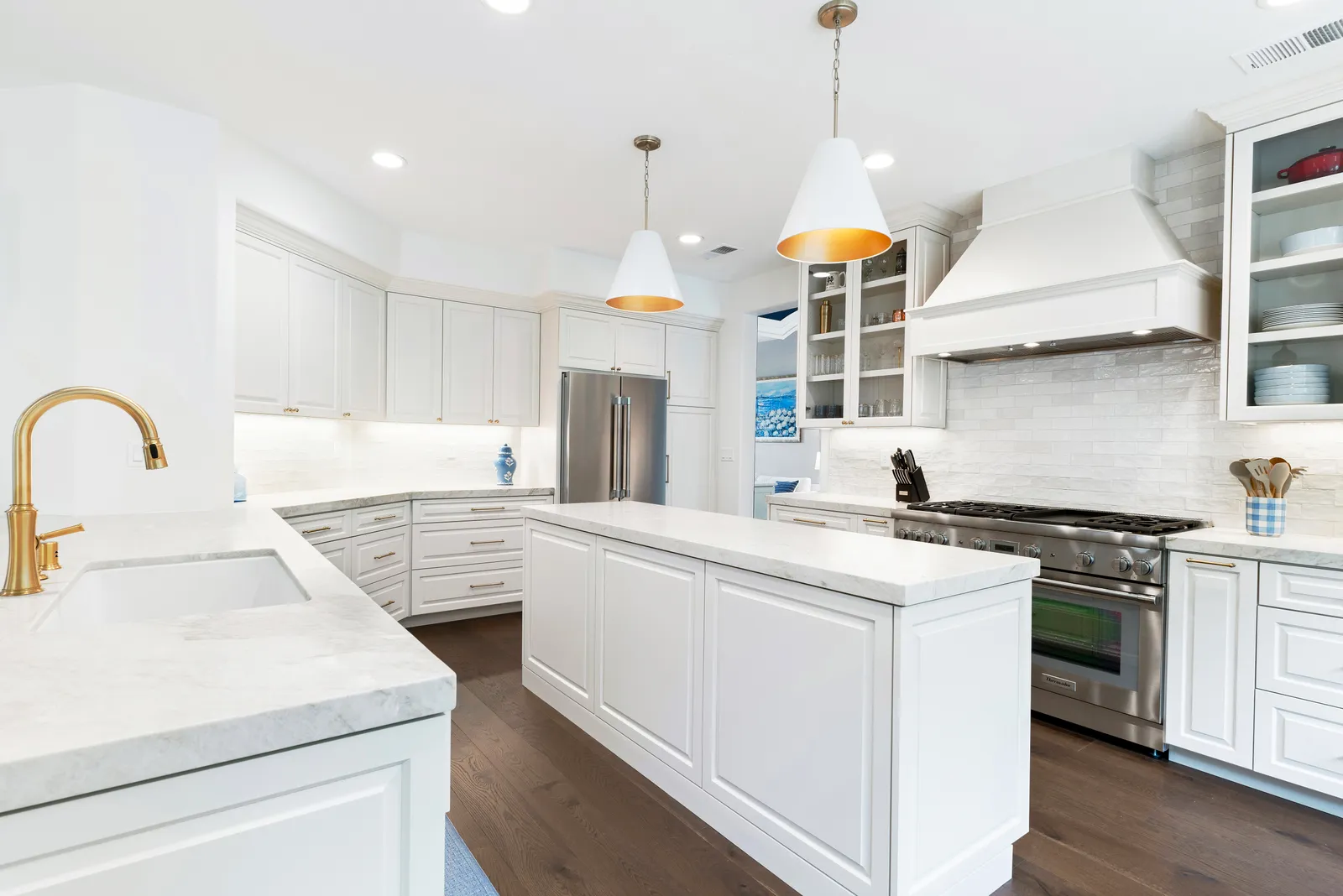 A white kitchen with marble counters, brass faucet and hardware, cone pendant lights, and a professional stainless range.