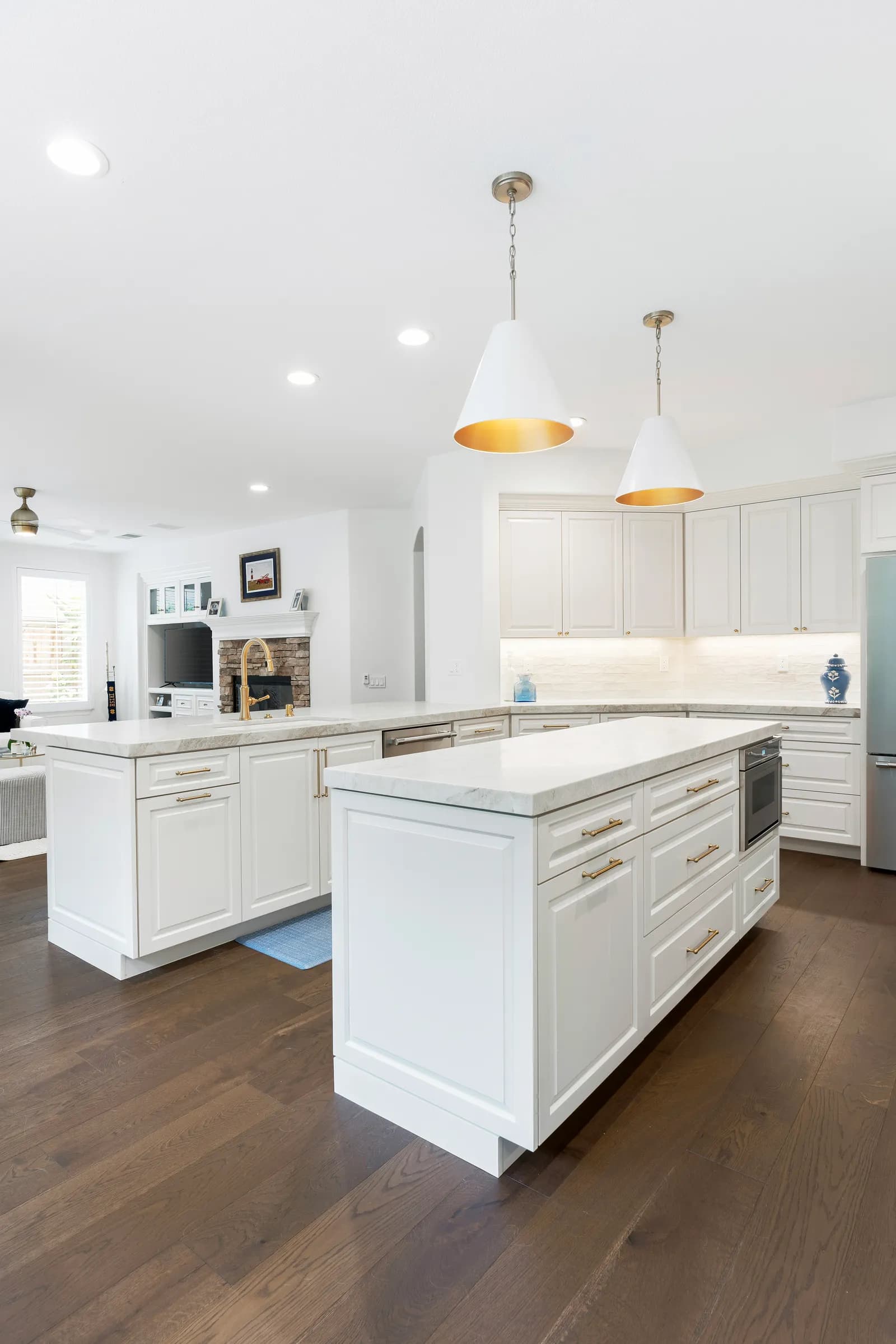 A bright white kitchen with a marble-topped island, brass pulls, cone pendants, and dark wood floors.
