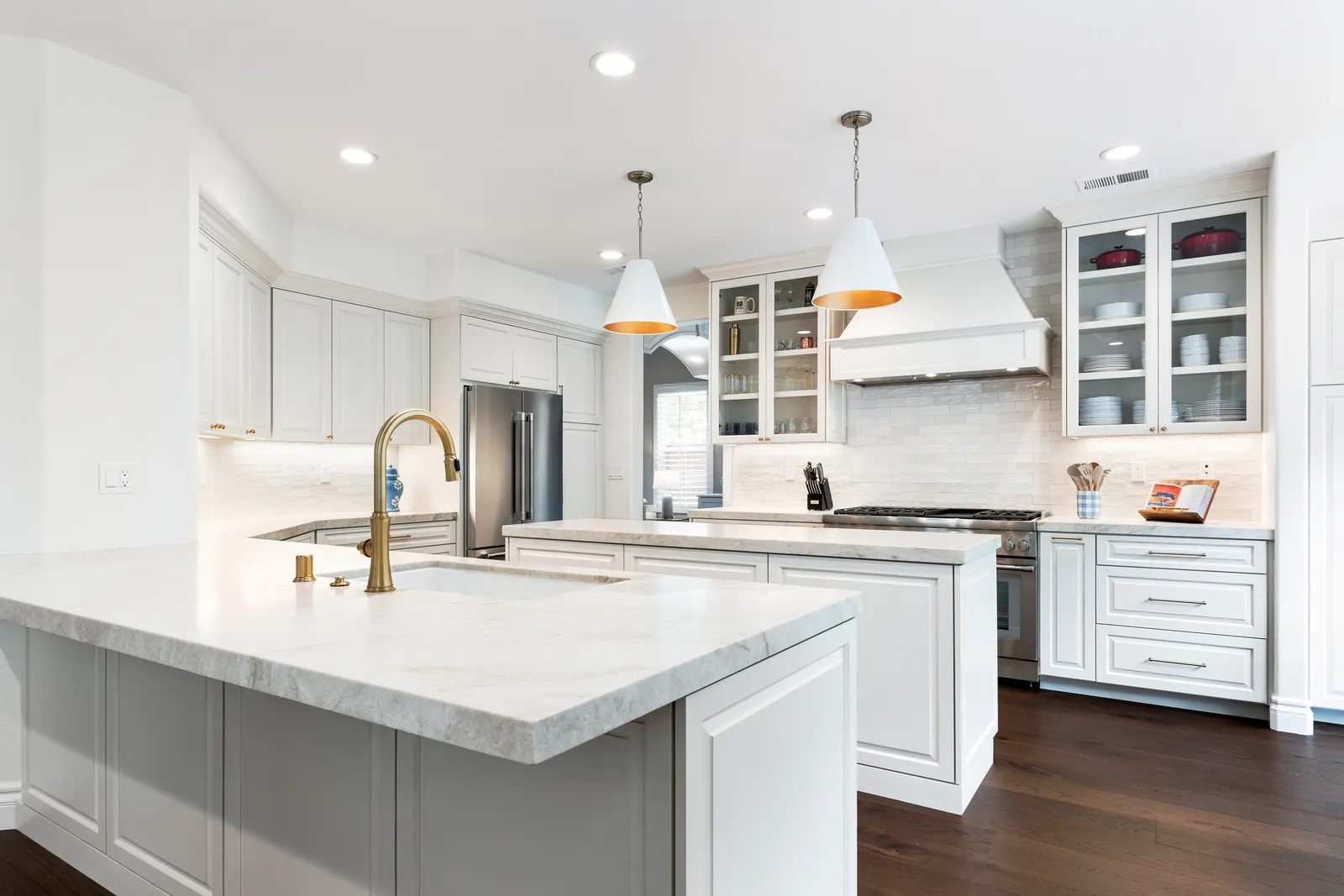 A white kitchen with a quartzite-topped peninsula, brass faucet, cone pendants, and dark hardwood floors.