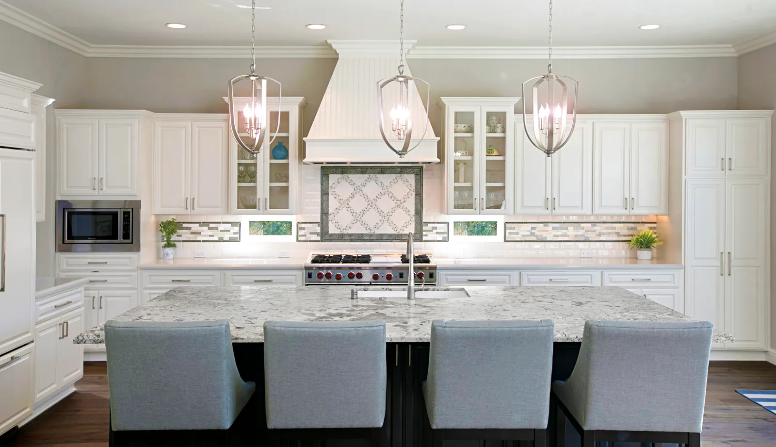 Traditional white kitchen with a long granite island, upholstered bar stools, and three lantern pendants.